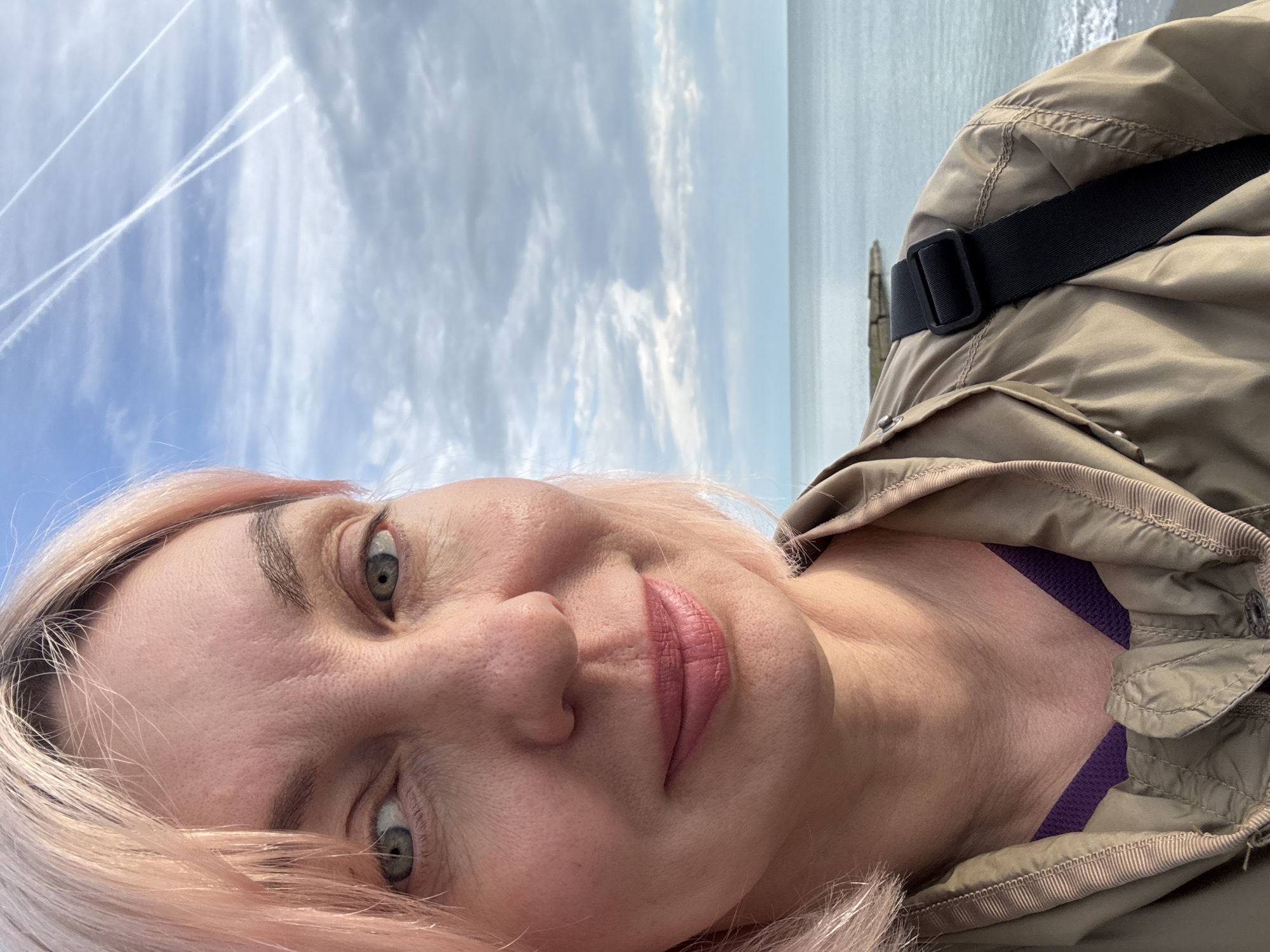 The image is a selfie taken outdoors with a clear blue sky with contrails and a body of water in the background. The subject is a woman with pink hair, wearing a khaki jacket over a purple top. She has green eyes and pink lipstick. A black strap is visible over her right shoulder. The environment suggests a coastal location. No evidence indicates it is Sochi, Russia.