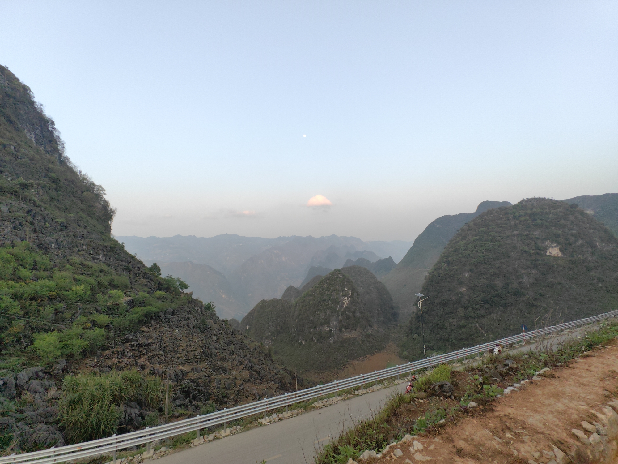 A scenic view in Đồng Văn Commune, Vietnam, features a winding road bordered by a metal guardrail on the right side. The foreground on the right is a reddish-brown, rocky terrain with sparse vegetation. In the middle ground, there are multiple layers of mountains and hills, with the closest ones covered in green vegetation. The distant mountains appear hazy due to the atmospheric perspective. A faint, light-colored object resembling the moon is visible in the pale sky. The sky has a slight gradient from a lighter color at the horizon to a slightly darker shade towards the top. The lighting suggests it may be early morning or late afternoon. A few small figures are visible on the road.