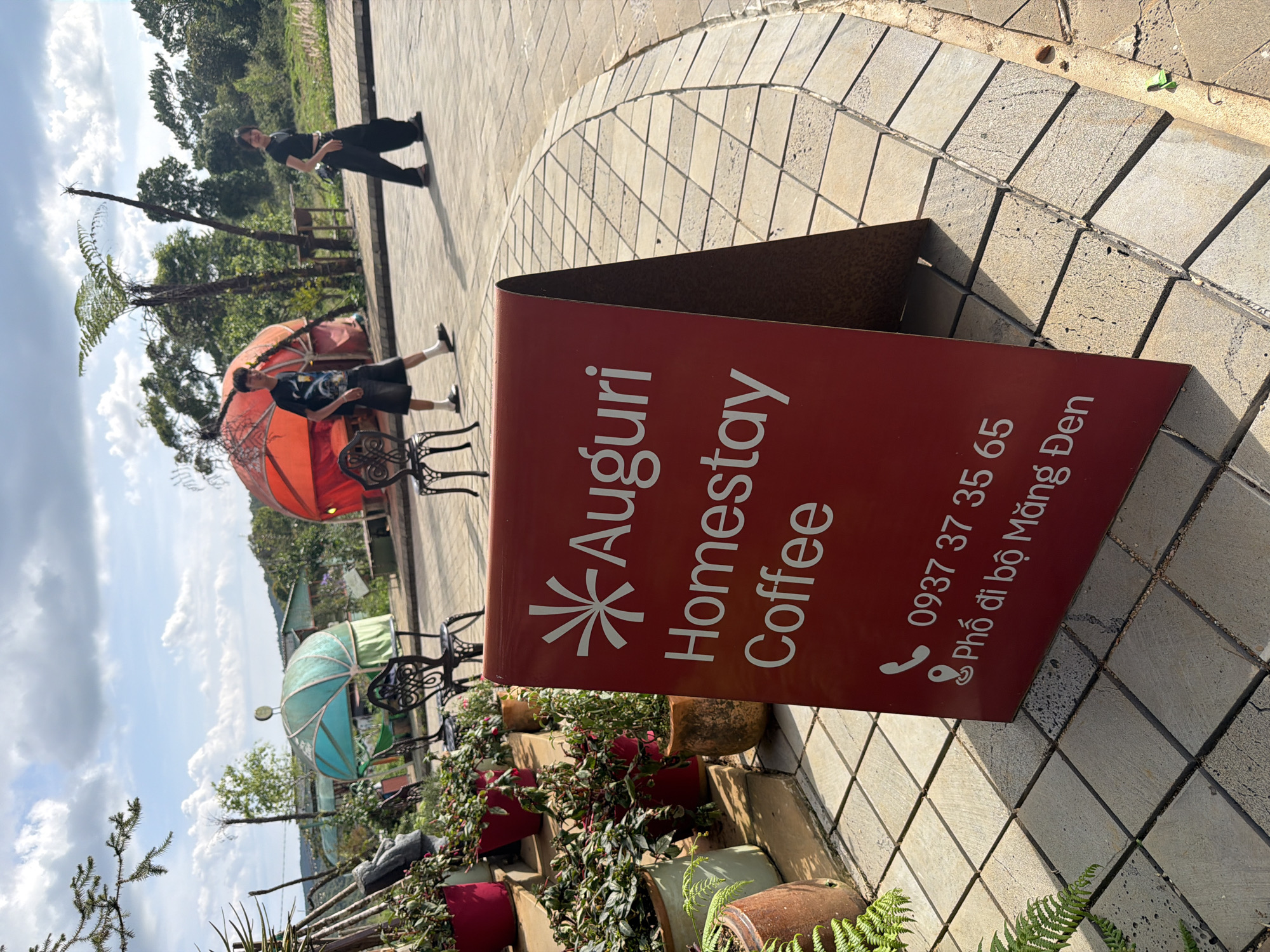 The image shows a red A-frame sign for "Auguri Homestay Coffee" located on a paved walkway in Măng Đen, Vietnam. The sign also displays a phone number (0937 37 35 65) and the phrase "Phố đi bộ Măng Đen," indicating the location is within the pedestrian area of Măng Đen. Behind the sign are planters with flowers. In the background, there are decorative umbrellas, benches, trees, and two individuals. One individual is standing near the umbrellas and benches, while the other is walking along the walkway in the distance. The weather appears sunny with a partially cloudy sky.