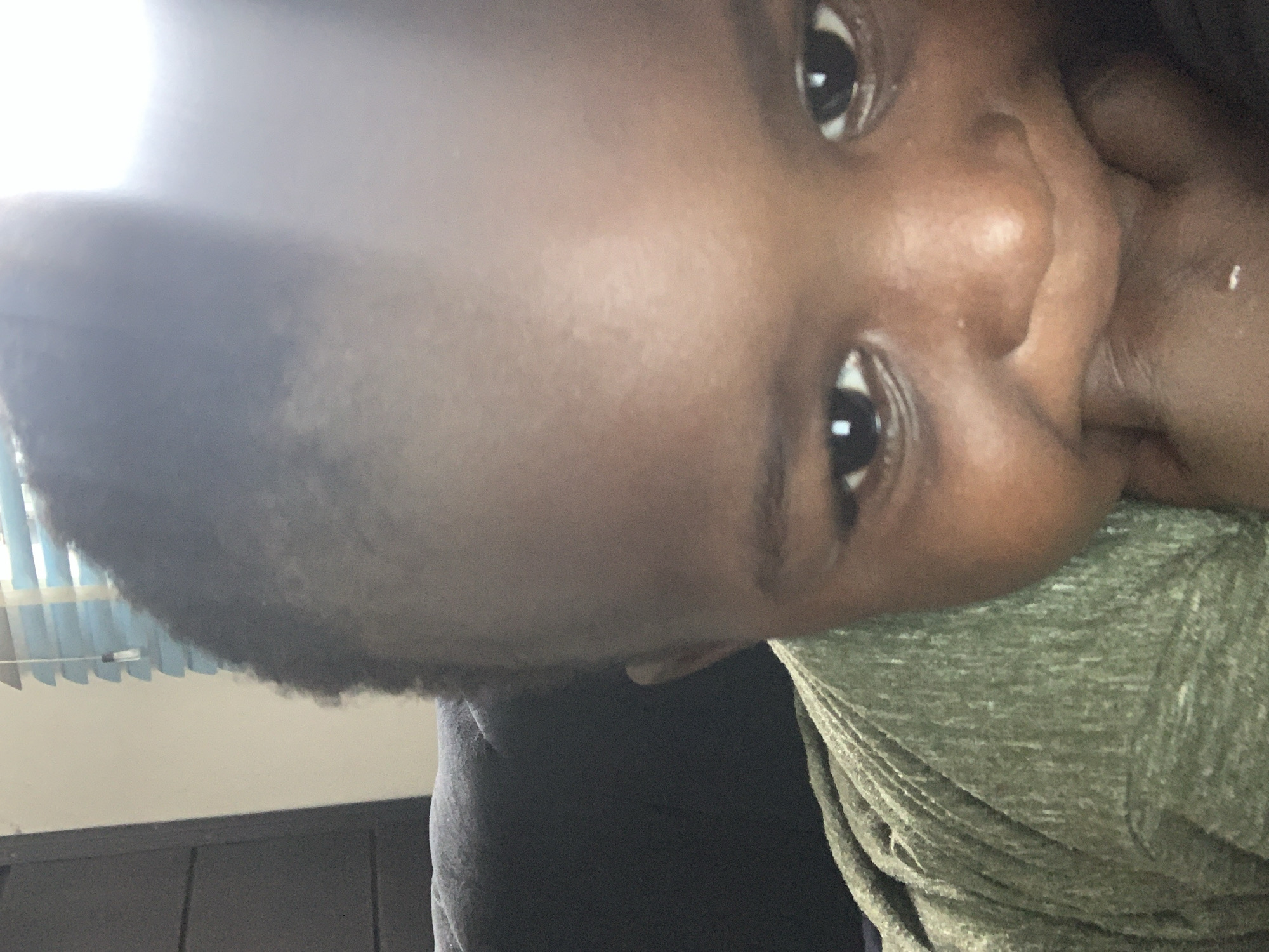 A close-up, head-and-shoulders shot of a young child is featured. The child has dark skin and curly hair. Their right eye is clearly visible, while the left eye is partially obscured by the chin. The child's mouth is covered by their hand. They are wearing a green, short-sleeved shirt. In the background, a dark blue cushioned chair is visible, as is a window with horizontal blinds. The lighting suggests an indoor setting.