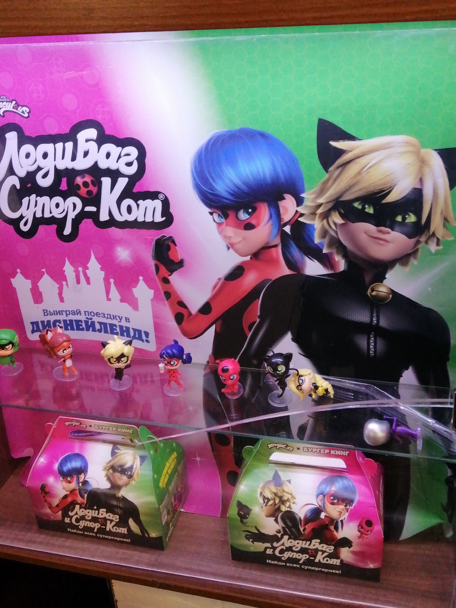 The image shows a display of "Miraculous Ladybug" merchandise, likely in a retail store. The display features a large backdrop with characters from the show, Ladybug and Cat Noir. Below this, on a glass shelf, are several small figurines of characters, including Ladybug, Cat Noir, and other characters from the series. In the foreground, there are two promotional boxes with images of Ladybug and Cat Noir and the text "ЛедиБаг и Супер-Кот" (Ladybug and Super-Cat) and "Найди всех супергероев!" (Find all the superheroes!). The boxes are shaped like small gift bags. The overall setting appears to be a store, possibly a toy store or a fast-food restaurant promotion, given the style of the boxes. The context of Rostov-on-Don, Russia, is not directly evident from the image itself but would likely be known from the location where the photo was taken.