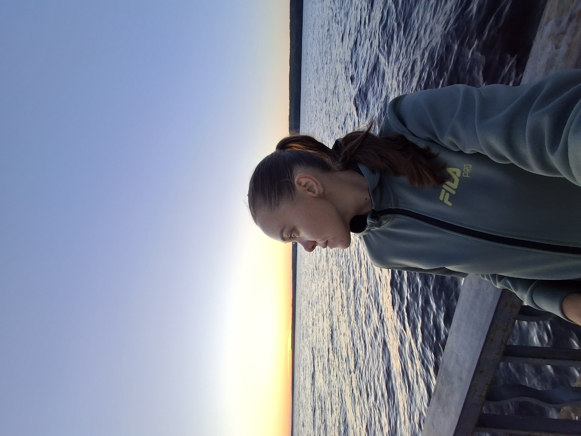 A woman is standing on a pier, looking out at a body of water during sunset. The water is calm with small ripples. The sky is clear and blue with a hint of orange and yellow from the setting sun on the horizon. The woman is wearing a light green hooded jacket with a zipper. Her hair is pulled back in a ponytail. The pier is made of metal and appears to be weathered. The location is the Kama River embankment in Chaikovsky, Russia.
