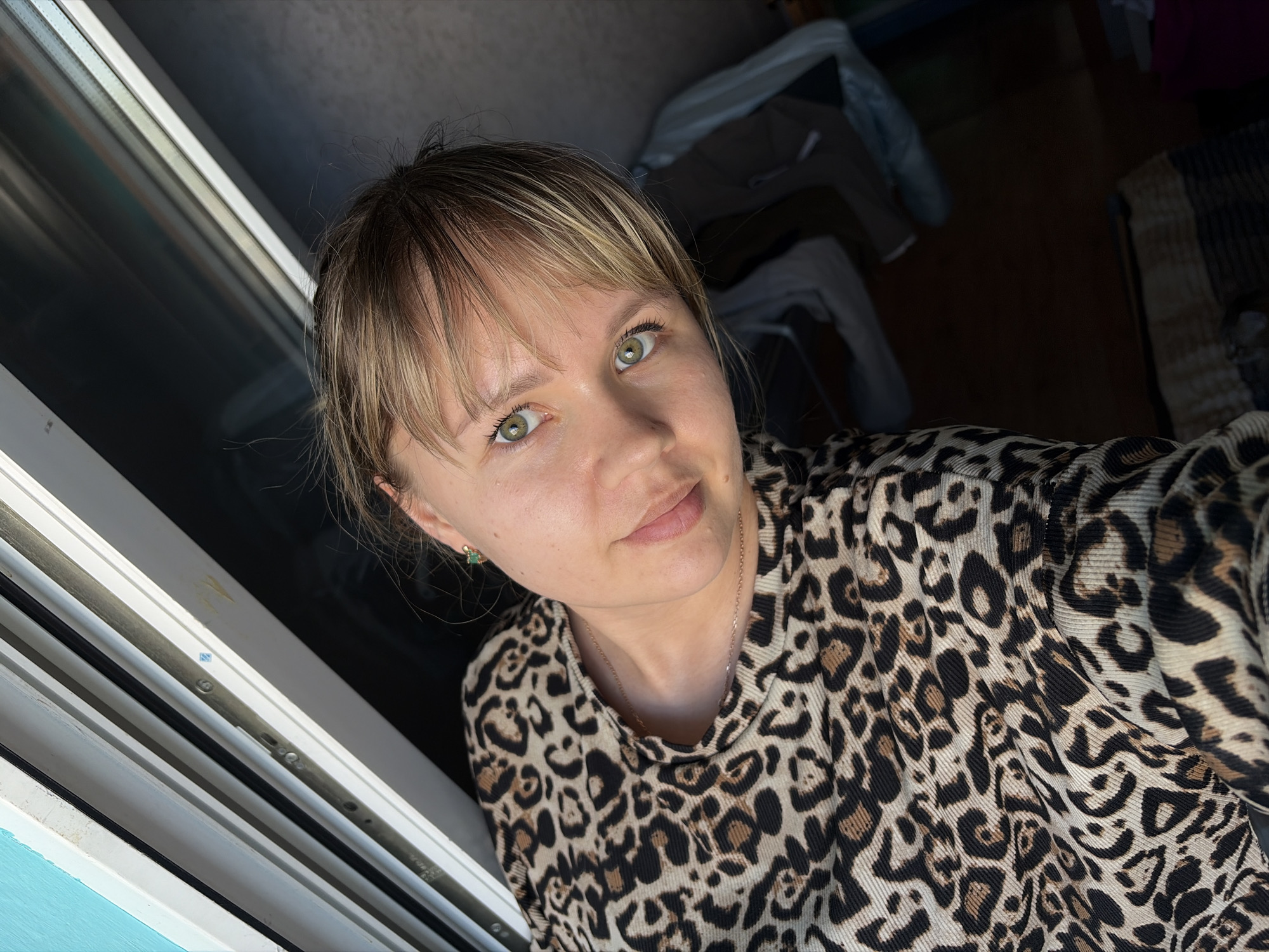A woman is posing for a selfie indoors, near a window. She has blonde hair, green eyes, and is wearing a leopard-print shirt. She is wearing a small, green earring and a thin chain around her neck. The window frame is white, and there is light coming in from the outside. In the background, clothes are draped over what appears to be a coat rack or clothes hanger, with a rug on the floor underneath. The location is in Chaikovsky, Russia.
