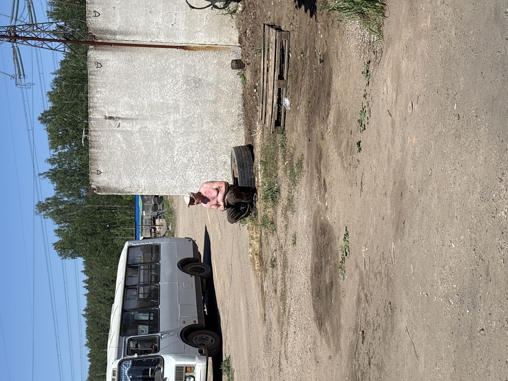 The image shows an outdoor scene with a white bus parked on a dusty surface in what appears to be an industrial or semi-urban area. A shirtless man with tan skin and a light-colored hat sits on a black tire next to a concrete wall. Behind the wall is a wooded area and electrical towers. To the side of the wall is an old wooden pallet. The location of the image is indicated as Noviy, Russia.