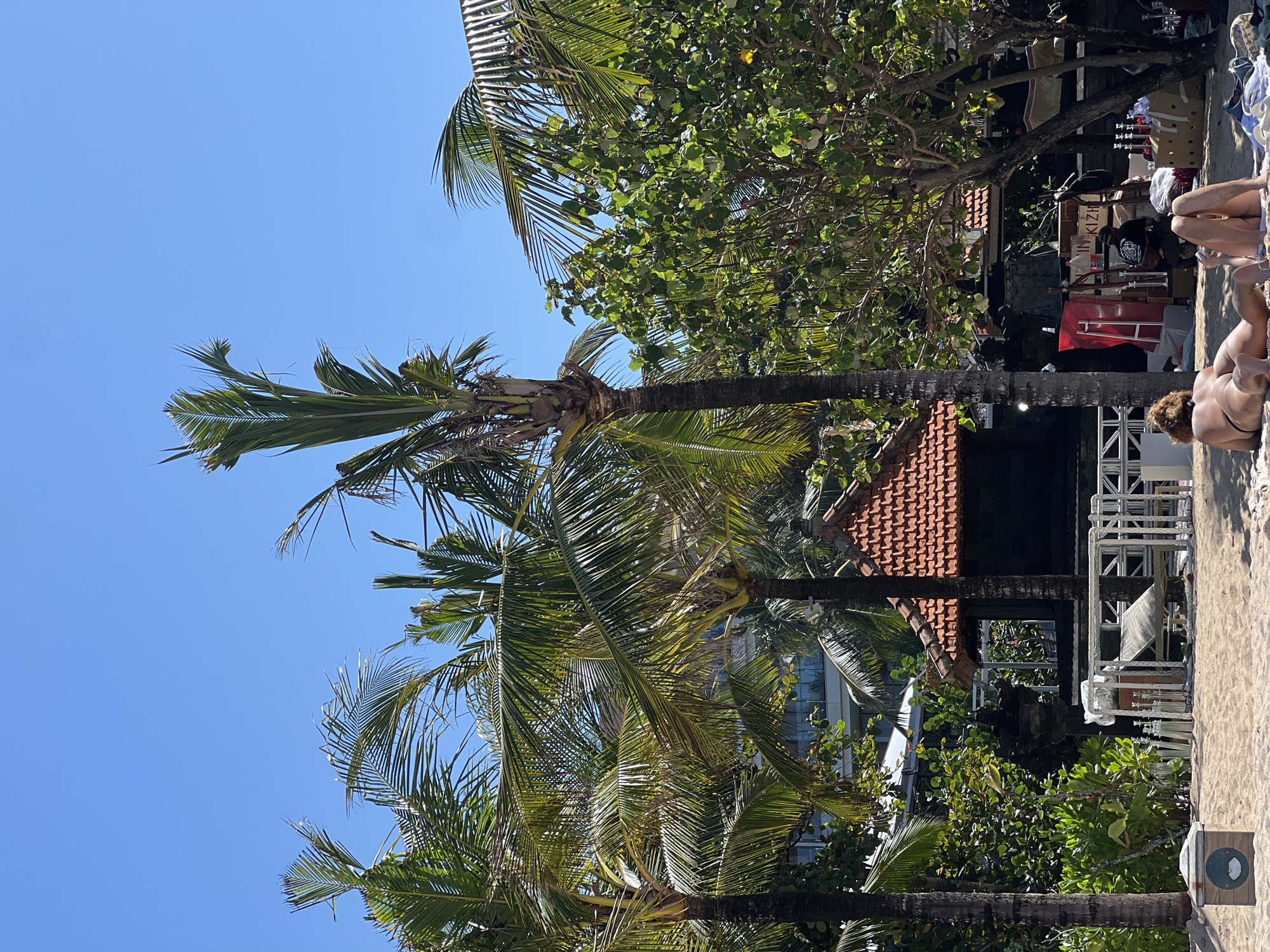 A sunny day at a beach in Kuta Selatan, Indonesia, with clear blue skies and lush green palm trees dominating the scene. In the foreground, two individuals are sunbathing on the sand. A small wooden structure with a red tiled roof is visible behind them, and further back, a modern hotel building can be seen amidst the tropical foliage. The overall atmosphere is one of relaxation and a tropical vacation.