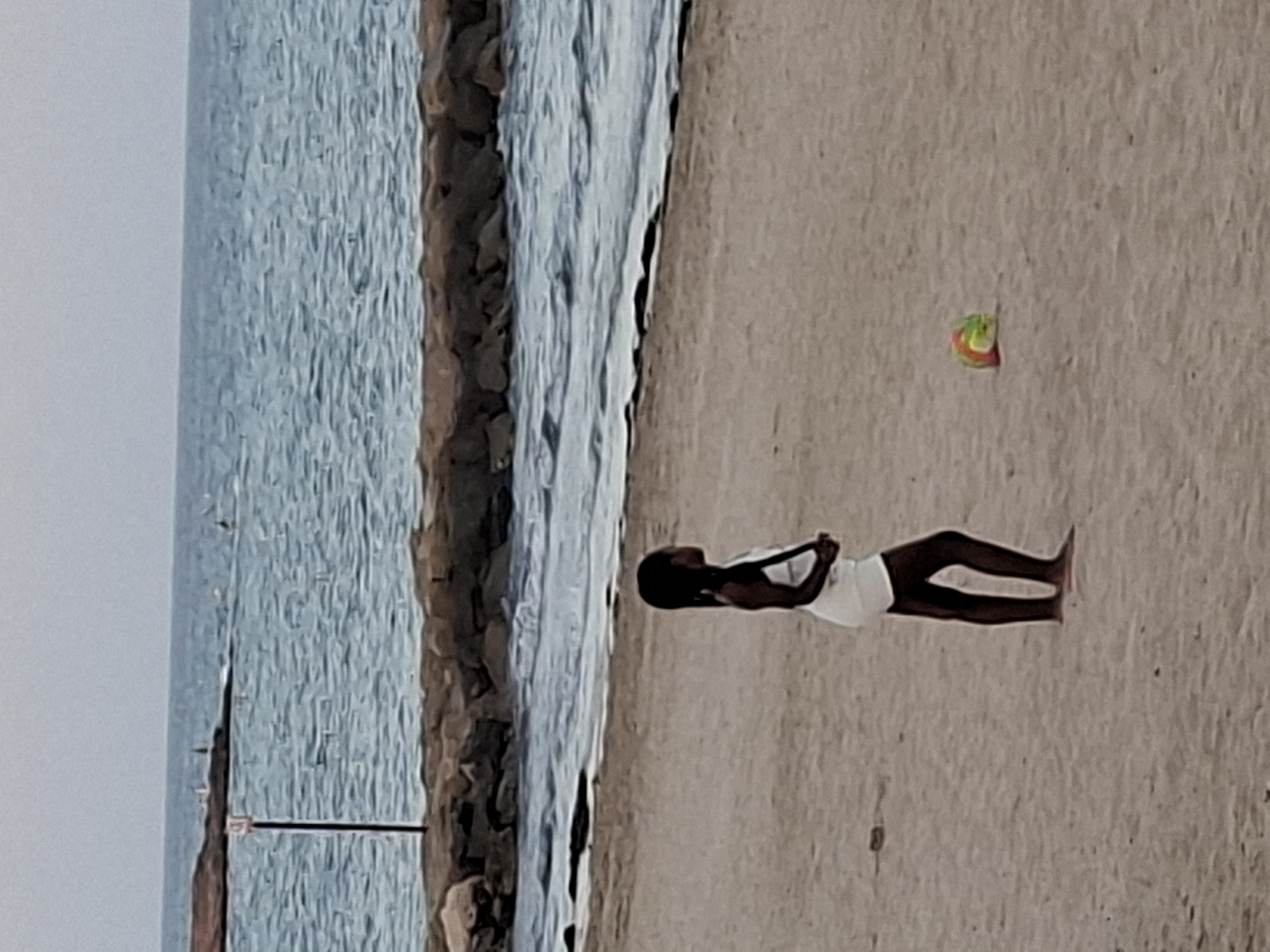 The image shows a full-length shot of a young Black girl standing on a sandy beach, with the ocean to her left. She is wearing a white tank top and black shorts. Her hair is pulled back in a bun, and she is looking towards the ocean. Her arms are bent at the elbows, and her hands are clasped behind her head.
The beach appears to be made of sand, and there are some rocks along the shoreline. The ocean is blue and calm, with small waves breaking on the shore. The sky is overcast, suggesting it may be a cloudy day. There is a small, colorful toy lying on the sand to the girl's right, possibly a bouncy ball with green, yellow, and red sections.
The scene suggests a relaxed and peaceful moment, possibly during a day at the beach. The girl's posture and the setting imply a moment of quiet reflection or enjoyment of the surroundings. The location context provided, Torrevieja, Spain, suggests a coastal town. There is no visible text in the image.