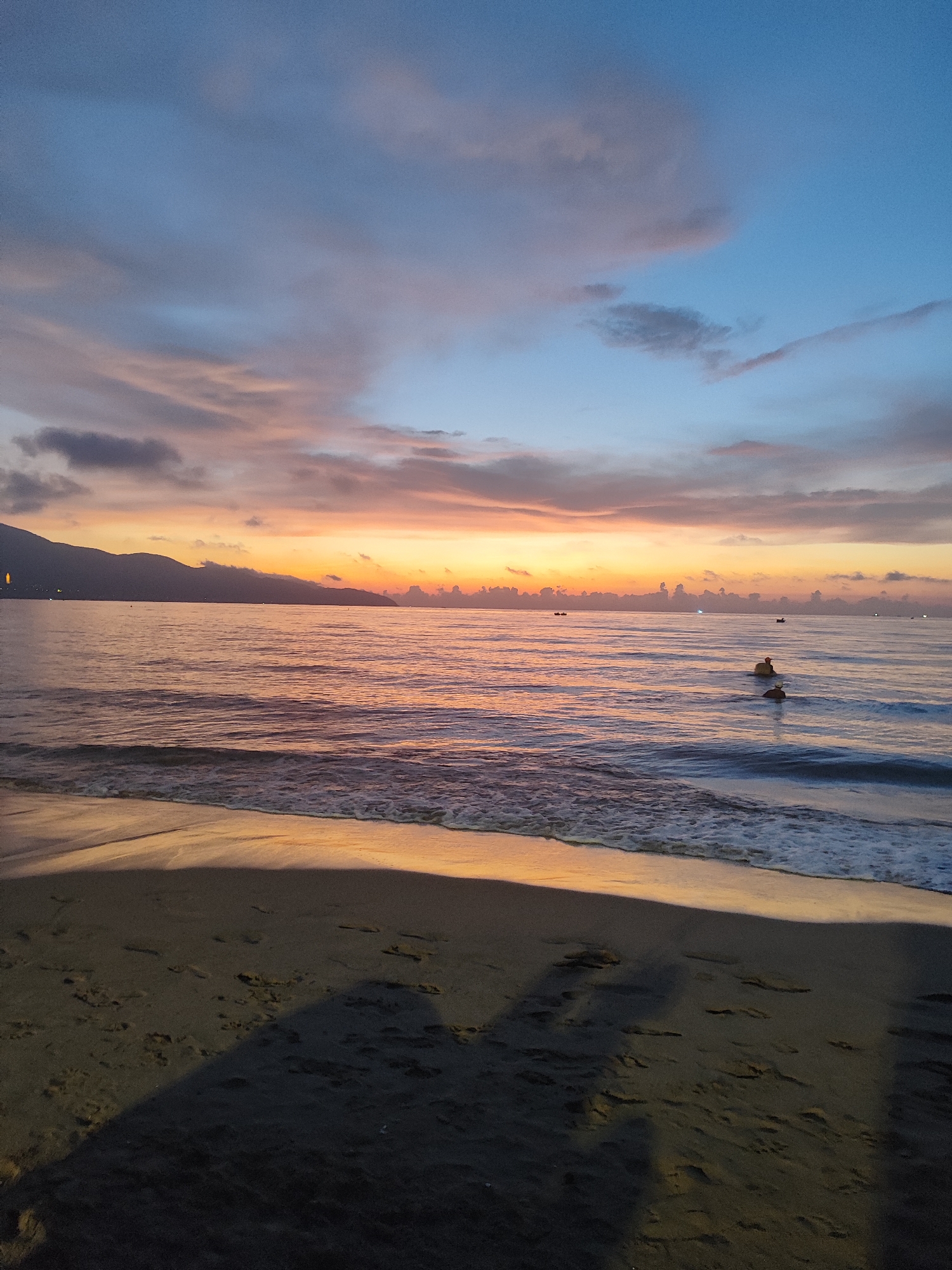 The image depicts a sunrise over the sea in Đà Nẵng, Vietnam. The sky transitions from blue to orange and pink hues with clouds. The calm sea reflects the colors of the sky, with gentle waves approaching a sandy beach. The silhouette of a mountain range is visible in the distance. On the beach, the shadow of two standing figures is visible. The beach is made of wet sand, while in the water, there are some barely visible distant boats.