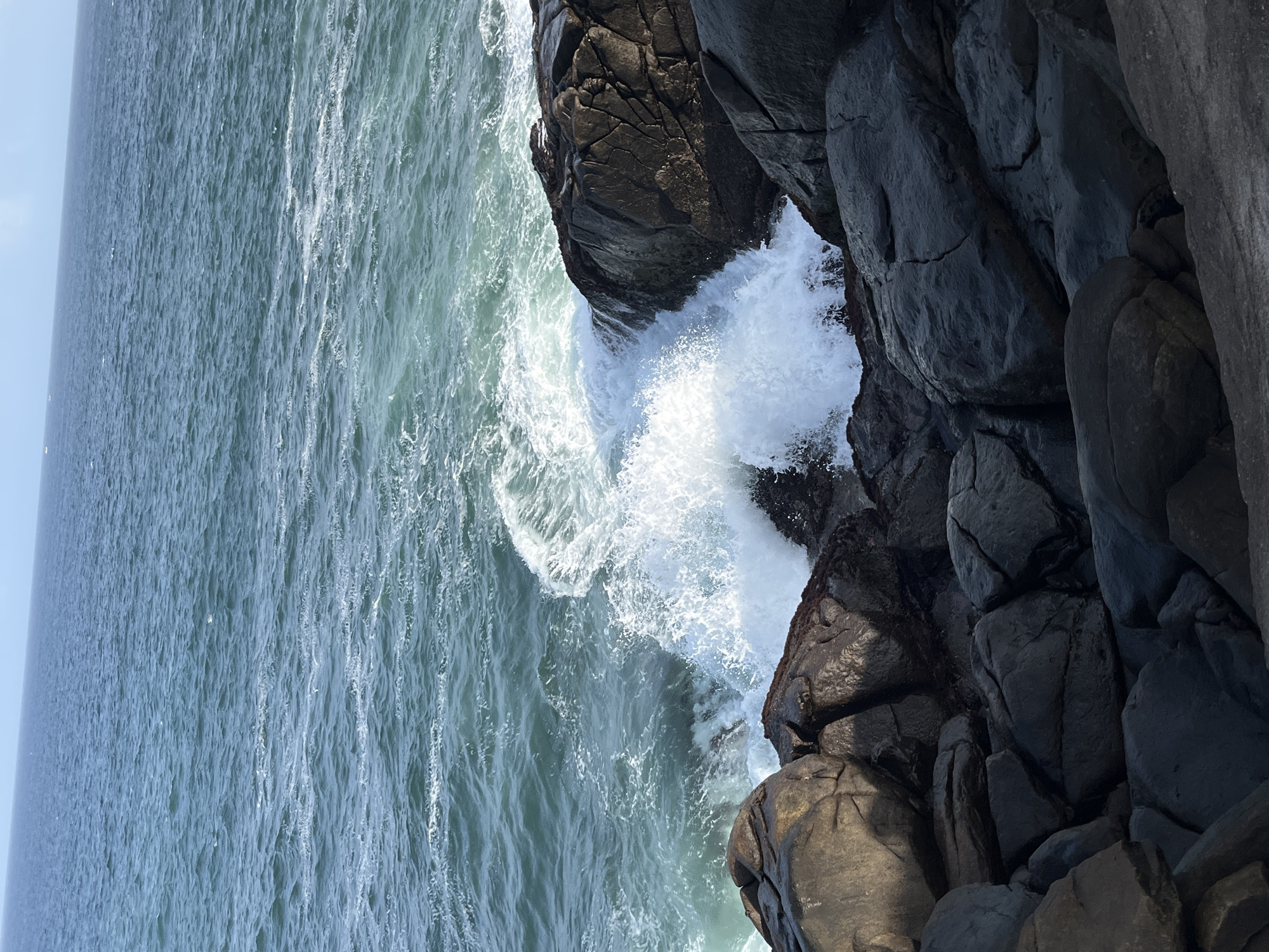 The image depicts a rugged coastal scene at Dondra, Sri Lanka, with dark, wet rocks dominating the foreground and midground. The rocks are varied in size and shape, some smooth and rounded, others angular and fractured. The sea is a deep teal, with the horizon line clearly defined against a pale sky. Waves crash against the rocks, creating white spray and foam that highlights the textures of the stone. The lighting suggests it is daytime, with the sun casting shadows that add depth and dimension to the rocky landscape. No people are visible in the frame.