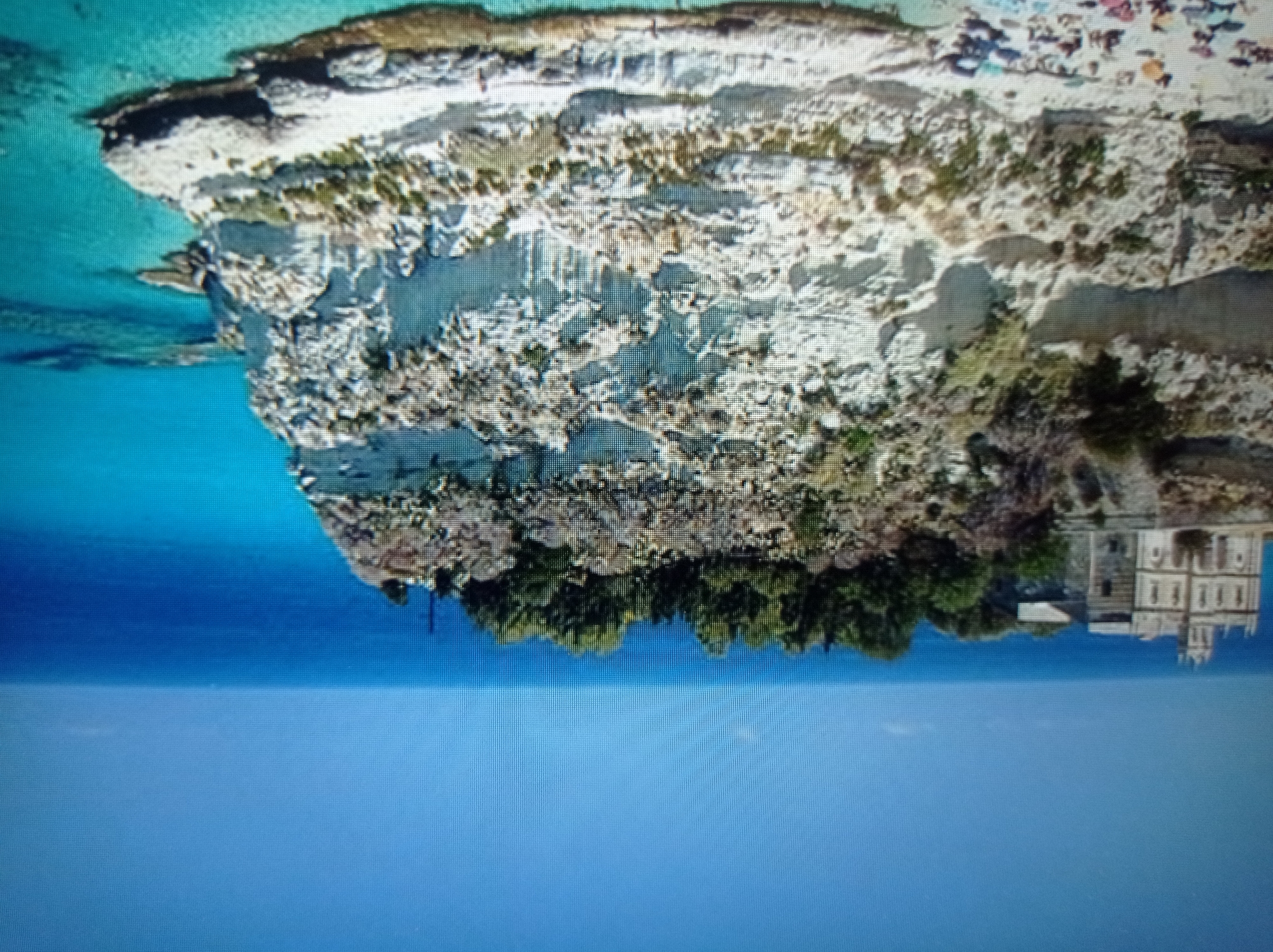 The image depicts a picturesque coastal scene dominated by a dramatic cliff face. On top of the cliff, a light-colored building with architectural details suggestive of a church or monastery stands prominently, surrounded by lush green trees. The cliff itself is composed of layered, pale rock with patches of vegetation clinging to its sides. At the base of the cliff, a sandy beach is visible, populated with numerous beach umbrellas and people, indicating a recreational area. The turquoise waters of the sea meet the shore, with clear visibility into the shallow areas revealing the seabed. The sky is a clear, vibrant blue, stretching to the horizon where it meets the sea. The overall impression is one of a serene and beautiful coastal landscape. The location is not in Mukachevo, Ukraine, but rather appears to be a Mediterranean coastal area, possibly Italy.