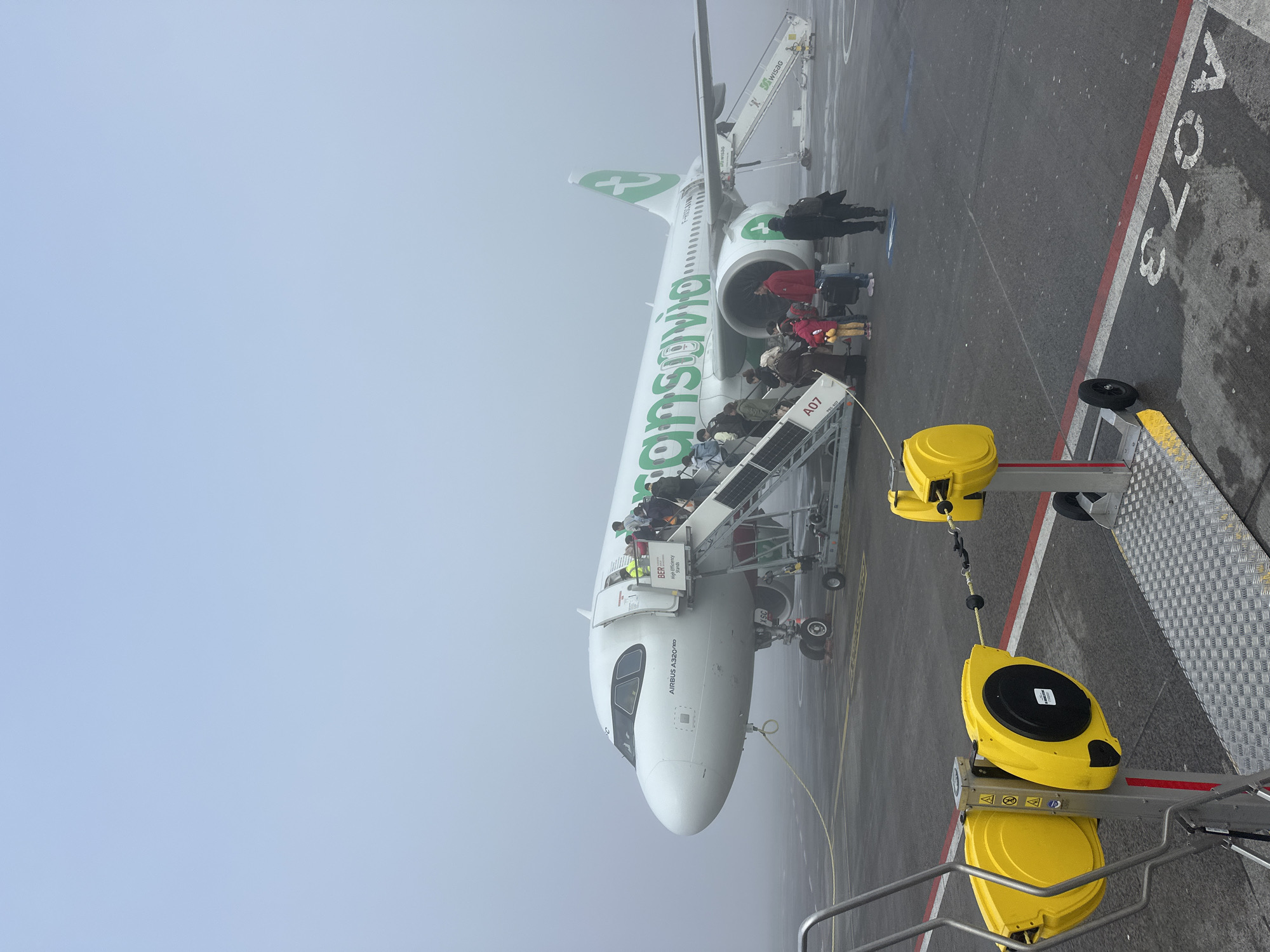 The image depicts a Transavia Airbus A320neo airplane being boarded by passengers via a mobile staircase at Berlin Brandenburg Airport (BER) in Schönefeld, Germany. The plane, with its distinctive green livery, is parked at gate A07. Passengers are ascending the stairs into the aircraft. A yellow safety barrier is attached to the mobile staircase. The image is taken in foggy or hazy conditions. The number "370A" is painted on the ground next to the plane.