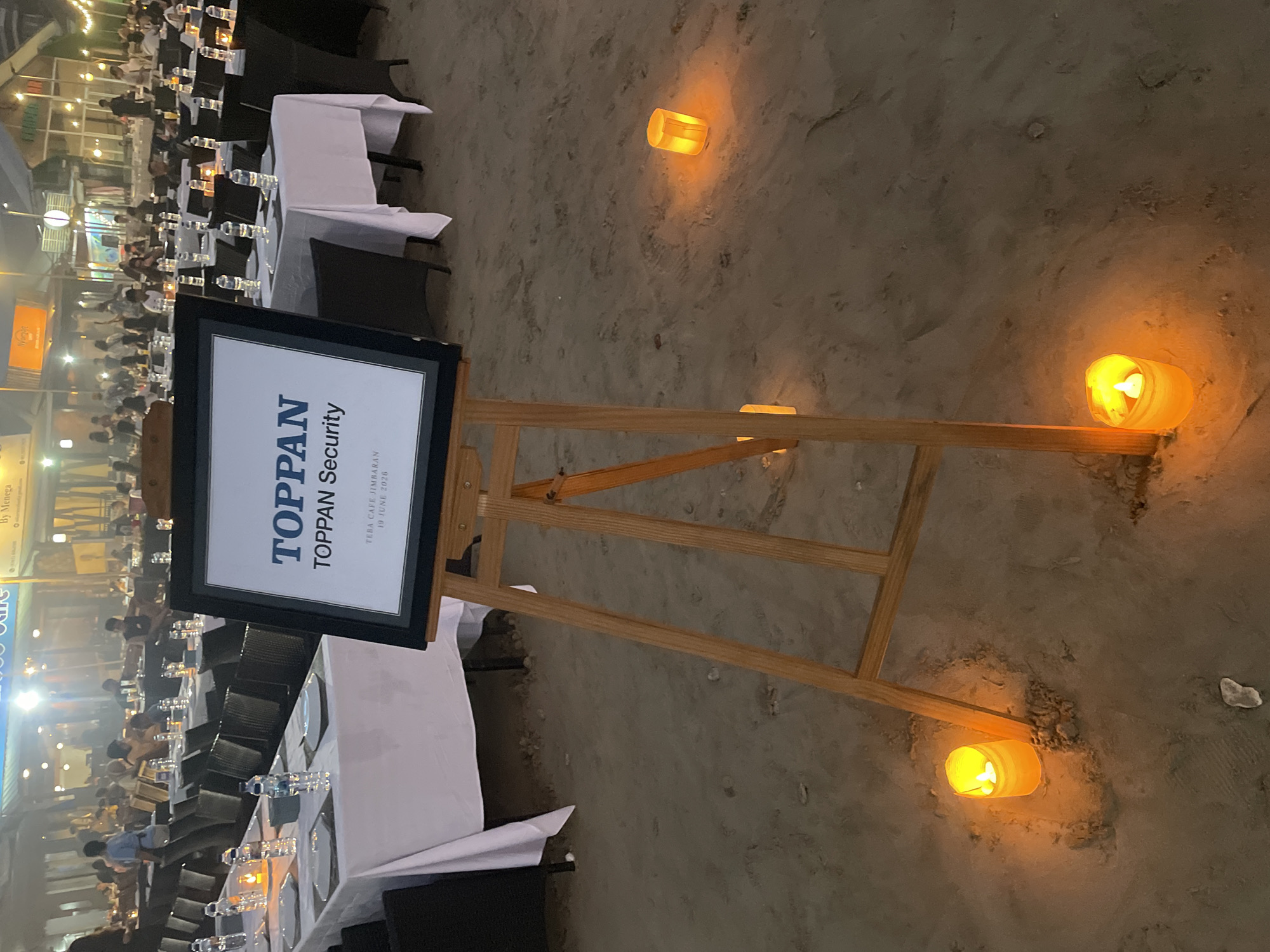 The image depicts an outdoor dining setup on a sandy beach at dusk or night. Several tables are covered with white tablecloths and set with plates and bottles of water. A large easel in the foreground displays a framed sign with the text "TOPPAN TOPPAN Security" and "TEBA CAFE JIMBARAN 19 JUNE 2020". Small, illuminated candles in orange holders are scattered on the sand around the tables and the easel, creating a warm, ambient glow. In the background, a row of buildings with lights on and a crowd of people are visible, suggesting a restaurant or event venue. The overall scene suggests a formal or semi-formal gathering, possibly a corporate event, taking place on a beach.