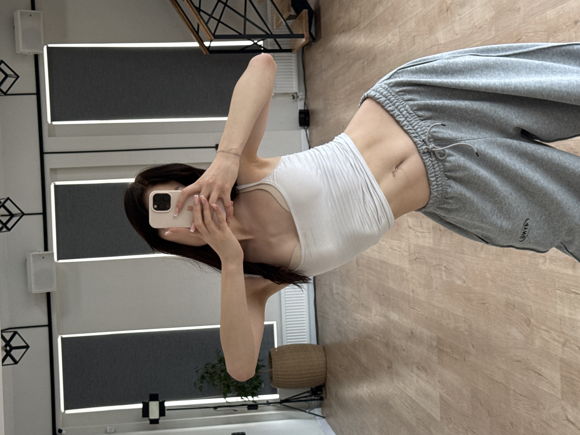 The image shows a young woman taking a mirror selfie. She is wearing a white ribbed tank top and grey sweatpants. The sweatpants have a drawstring and a small logo on the left leg which appears to say "(DMYK)". She has a belly button piercing. The background is a studio or dance space with wooden floors and a large mirror reflecting the room. There are black framed panels with white light strips on the wall behind her. A woven basket and a plant are also visible in the reflection. The lighting suggests it is daytime. The overall mood is casual and relaxed.