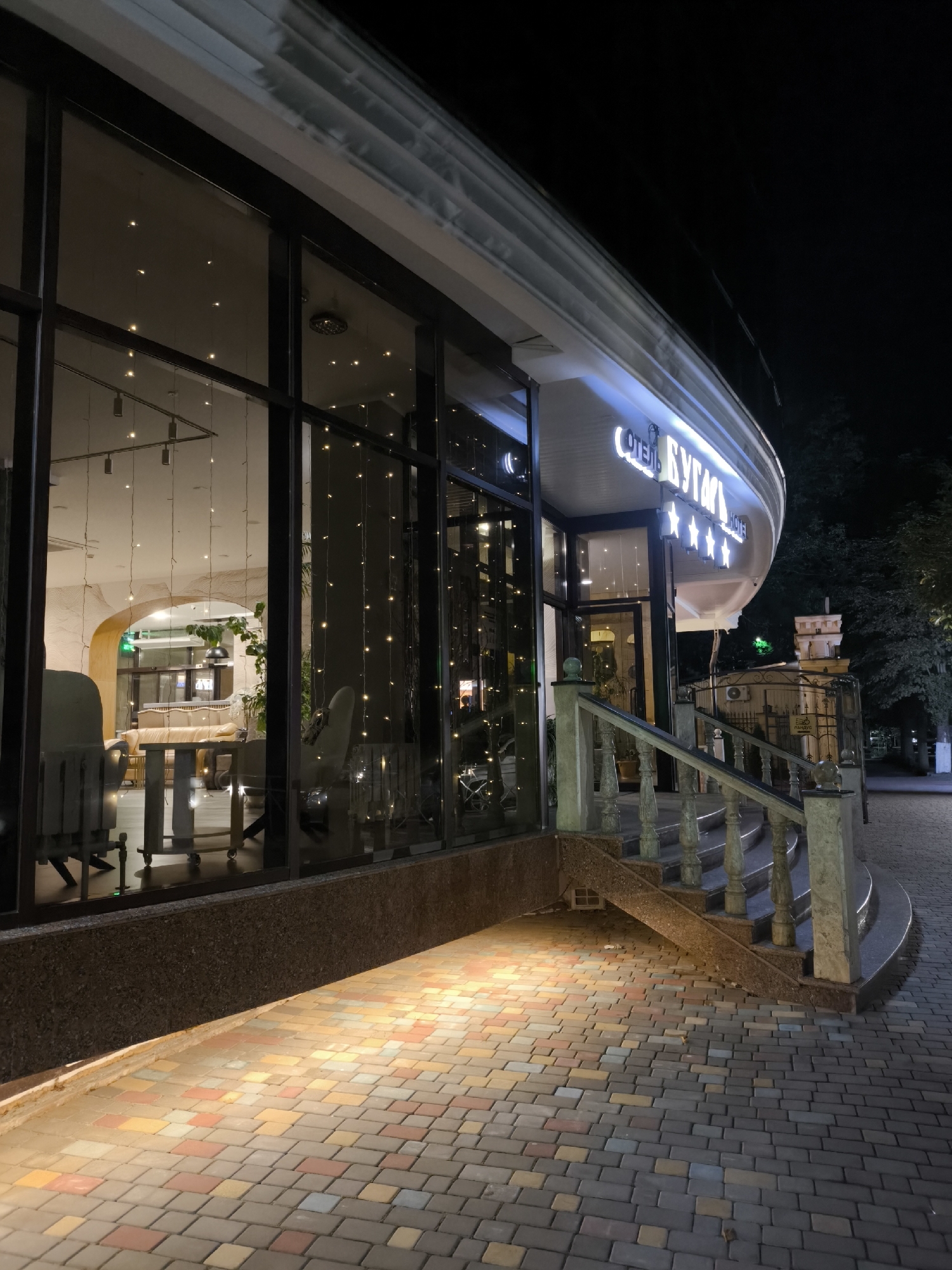 The image captures the exterior of the "ОТЕЛЬ БУГАЧ" (Hotel Bugach) in Yessentuki, Russia, at night. The hotel features large, reflective windows showcasing an interior decorated with string lights, illuminated furniture, and architectural elements. A curved staircase with ornate balustrades leads up to the entrance, with a patterned brick pathway illuminated by warm lighting. The hotel's name is prominently displayed in glowing letters above the entrance. In the background, a small, decorative tower and trees are visible. There are no people present in the scene.
