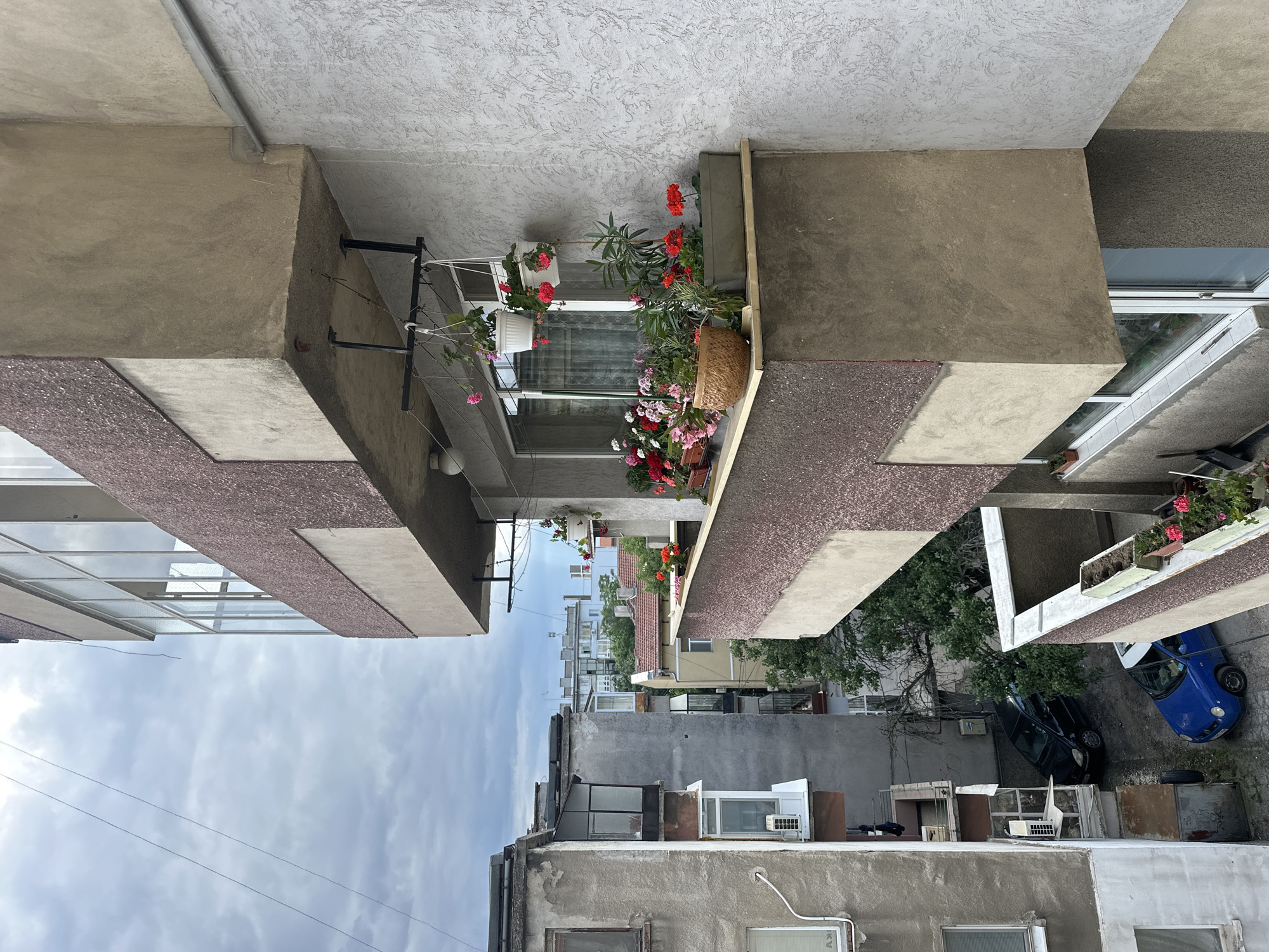 The image shows a view from a balcony in Ruse, Bulgaria, looking down at adjacent buildings and a narrow street. The foreground features a concrete balcony with several potted plants, including red and pink geraniums and other greenery. The balcony itself is decorated with a textured brown facade.
Below the balcony, a narrow, paved street is visible, with two cars parked: a dark-colored sedan and a smaller blue car. The buildings surrounding the street are multi-story apartment buildings with various architectural styles and states of repair. Some balconies are enclosed with glass, while others are open. The sky is overcast with gray clouds. The overall scene depicts a residential urban environment.