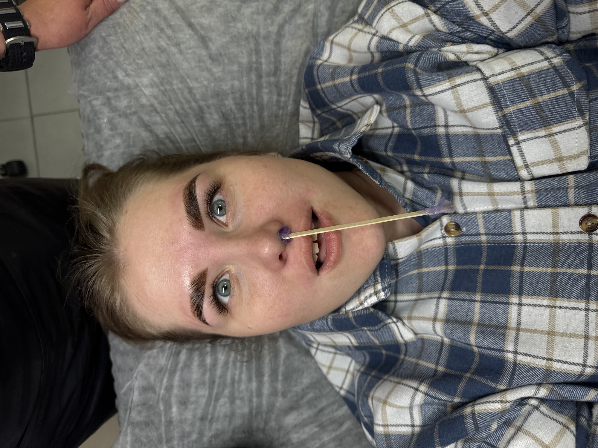 A young, fair-skinned woman with light blue eyes and recently groomed eyebrows is lying on a gray sheet or blanket. She is wearing a blue and white plaid shirt that is partly unbuttoned. A wooden stick with a small amount of purple wax on the end is placed between her nose and upper lip. A hand wearing a black watch is visible in the upper left corner of the image. The location is likely a beauty salon in Perm, Russia, based on the assumption of the context. The woman is likely undergoing a facial hair removal procedure.