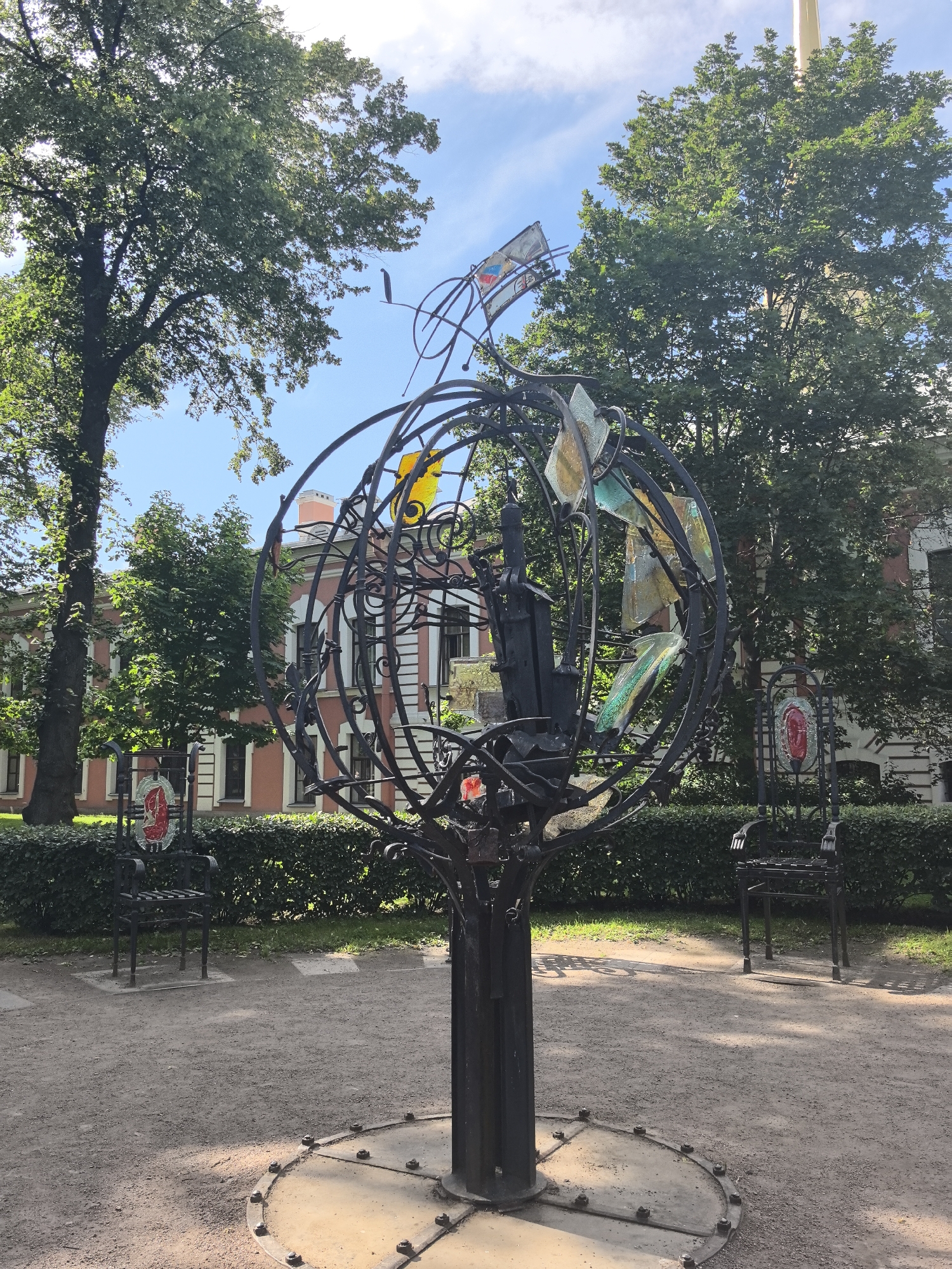 This image captures an outdoor scene featuring a unique, large-scale metal sculpture that resembles a globe or a celestial sphere. The sculpture is intricate, made of dark, wrought metal, with a cylindrical base anchored to a circular metal plate embedded in the ground. Within the sphere, various pieces of colorful, translucent glass are embedded, adding a vibrant contrast to the dark metal structure. Surrounding the sculpture, the ground is covered with gravel, and a manicured hedge forms a backdrop.
In front of the hedge and flanking the sculpture, two identical, ornate metal chairs are visible. These chairs are also made of dark metal and appear to be part of the artistic installation, perhaps inviting viewers to sit and contemplate the sculpture. Behind the hedge and chairs, a light pink building with white trim and numerous windows is partially visible through the lush green foliage of tall trees. The building's architecture suggests a historical or institutional setting.
The weather appears to be pleasant, with a bright blue sky dotted with fluffy white clouds. The sun is shining, casting dappled shadows from the trees and the sculpture onto the gravel path, indicating it is likely daytime, possibly late morning or early afternoon. The overall atmosphere is serene and artistic, suggesting a public park or a garden setting in Saint Petersburg, Russia, dedicated to art and public enjoyment. No people are visible in the frame, and there is no discernible text.