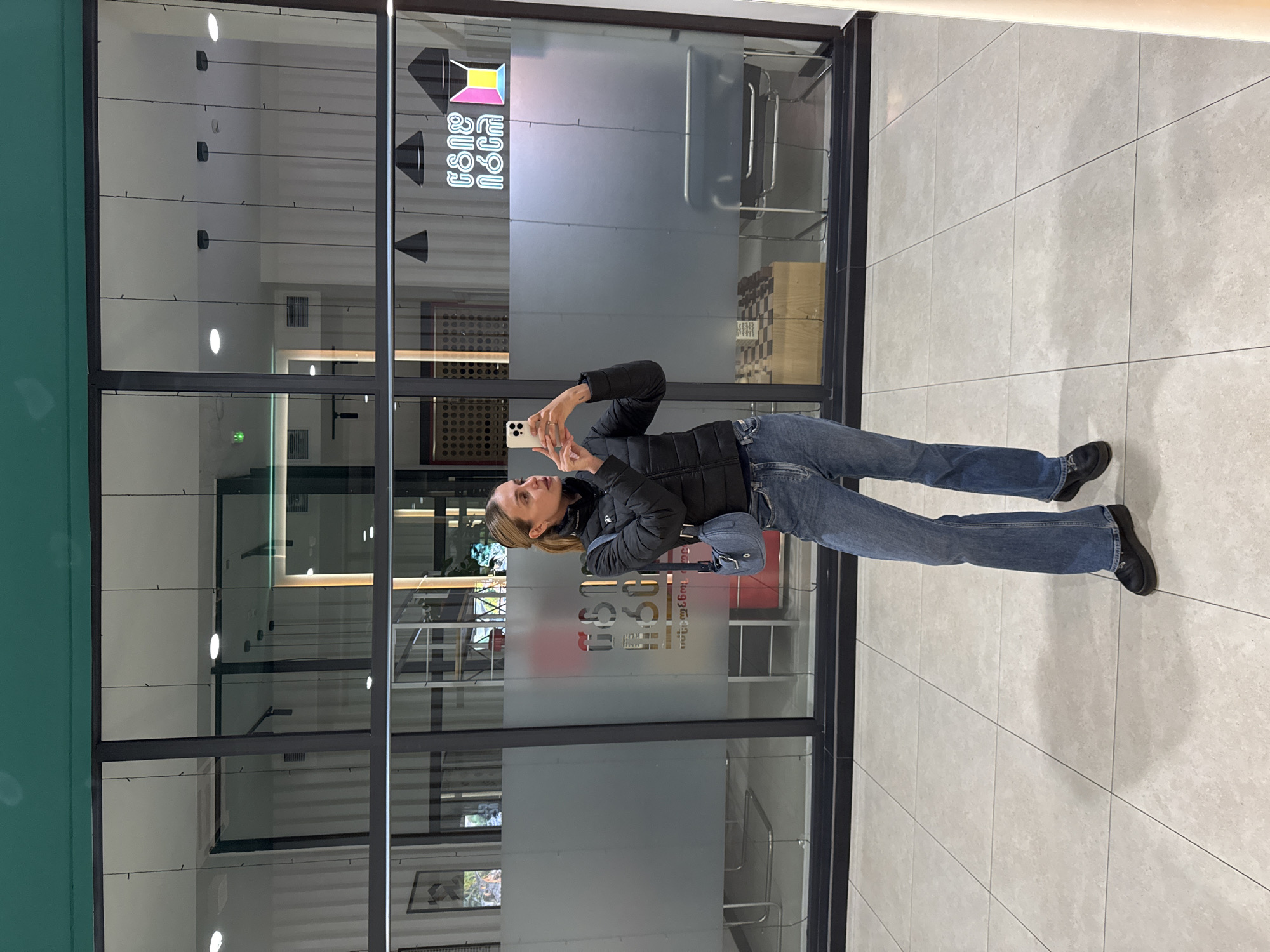 A woman in a black jacket and jeans is standing and taking a picture with her phone in front of a glass window with a logo and Georgian text that reads "თბილისი ქუჩები Tbilisi Streets". She has a small blue purse slung over her shoulder. The background includes an interior with various furniture elements and the exterior floor which is tiled. The location is likely in Tbilisi, Georgia.