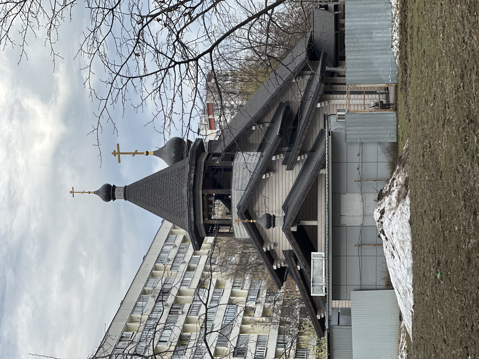 The image depicts a wooden church in Moscow, Russia. The church has a dark gray roof and a spire topped with a golden cross. Adjacent to the church is a tall, modern apartment building with many windows. Construction or maintenance work is underway around the church, as evidenced by temporary fencing and metal sheeting. There is a patch of melting snow next to the church, and a bare tree branch is visible in the foreground against a cloudy sky.