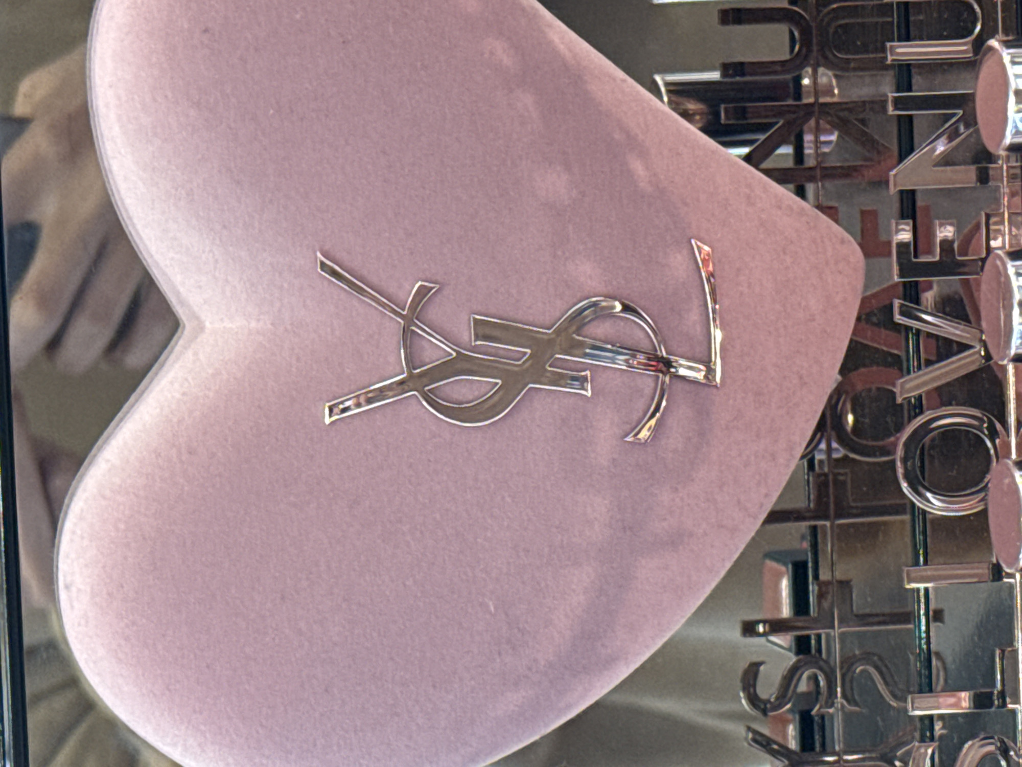 A close-up, eye-level shot shows a pale pink, heart-shaped object with the silver Yves Saint Laurent logo embossed on it. The object appears to be made of velvet. Below the heart, metallic letters spell out "VENUE" and "S L O V E N U". The background is blurred, but suggests a display or retail environment, possibly in a mall. The scene is well-lit, with reflections visible on the metallic letters.