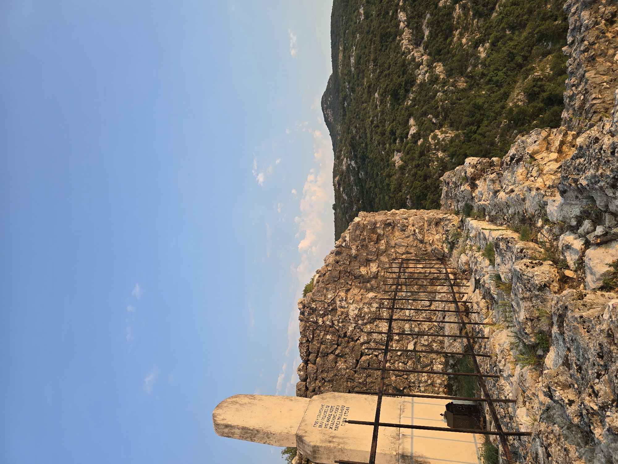 A tombstone and crumbling stone wall stand on a rocky outcrop overlooking a densely forested hillside under a clear blue sky with scattered clouds. The tombstone is a weathered, light-colored obelisk with inscriptions in Cyrillic script. A black metal fence surrounds a portion of the area in front of the tombstone and wall, and a small, dark metal lantern hangs on the fence. The landscape beyond the immediate site is characterized by rolling hills covered in lush green vegetation, with a single prominent peak in the distance. The lighting suggests it is late afternoon or early evening, with warm sunlight illuminating the rocks and vegetation. The scene appears to be a historical or memorial site, likely located in the mountainous terrain of Gornja Vrbica, Montenegro.