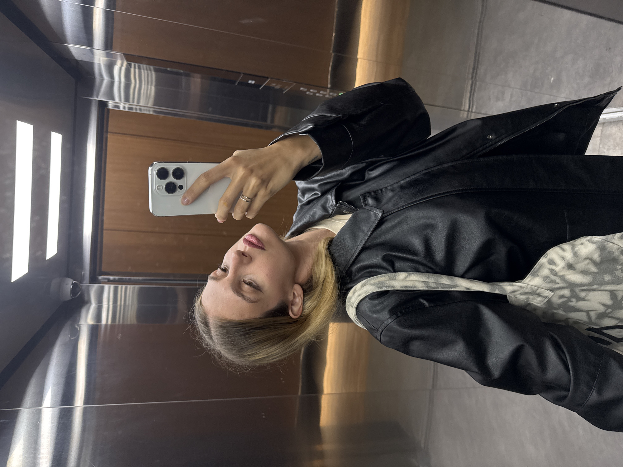A woman in a black leather jacket is taking a selfie in an elevator. She is holding a white smartphone in her right hand. The elevator has metal walls and wood paneling. Three vertical lights are visible on the left wall. The location is in the city of Unknown, Unknown.