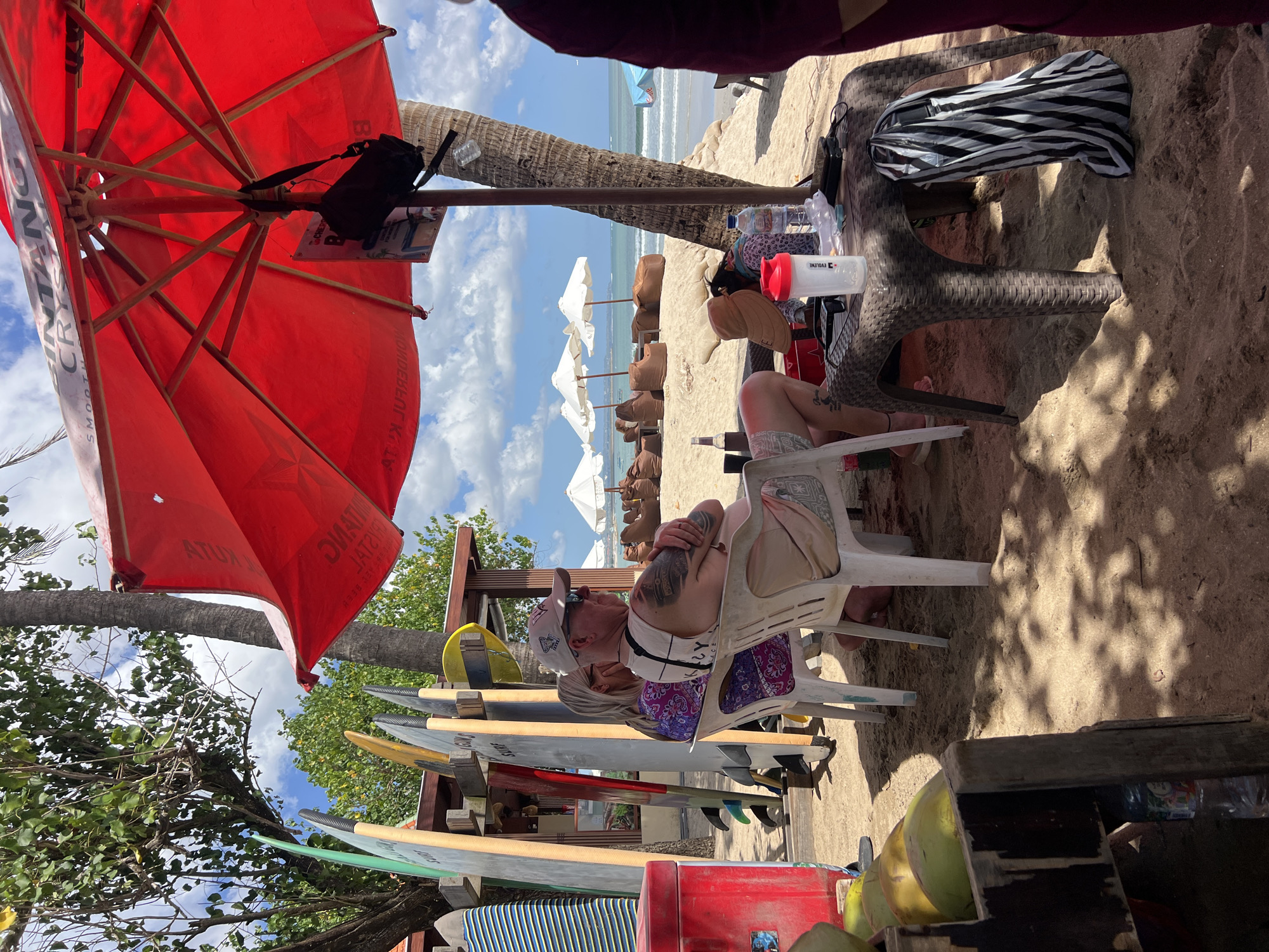In Kuta, Indonesia, on a sandy beach, a group of people are relaxing under a large red umbrella. The umbrella is branded with the text "Bintang Crystal Smooth". In the foreground, several surfboards are propped up, some leaning against a wooden structure. A red cooler is partially visible to the left, filled with green coconuts.
Two individuals are seated on white plastic chairs in the midground. The man, wearing a baseball cap and sunglasses, has his arms crossed and appears to be looking out towards the ocean. He has tattoos on his arms. The woman next to him is wearing a purple and pink patterned top.
A brown woven plastic table is positioned in front of the seated individuals, holding a white shaker bottle, a water bottle, and a black electronic device. A striped bag hangs from the table.
In the background, rows of brown beanbag chairs are arranged on the sand, with white beach umbrellas providing shade. The ocean is visible with gentle waves, and a blue umbrella stands further down the beach. Palm trees and lush green foliage frame the scene. The overall atmosphere suggests a casual, tropical beach setting.