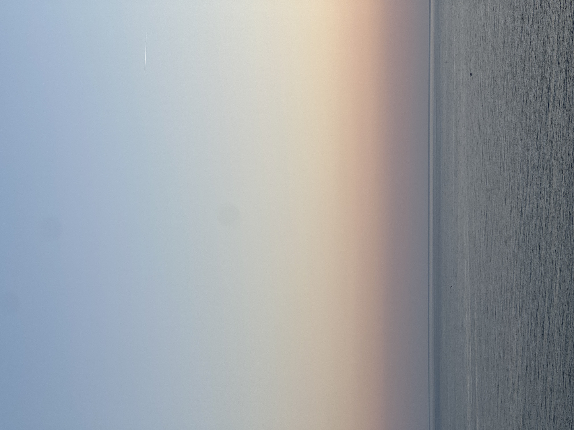 The image depicts a seascape at dusk or dawn. The left side of the frame shows a gradient sky shifting from blue to a pale peach color. On the right, the sea extends into the horizon, marked by vertical lines of light reflecting on the water surface. There are two small objects or vessels visible in the sea. A faint contrail appears in the upper left of the sky. The location is possibly Šušanj, Montenegro.