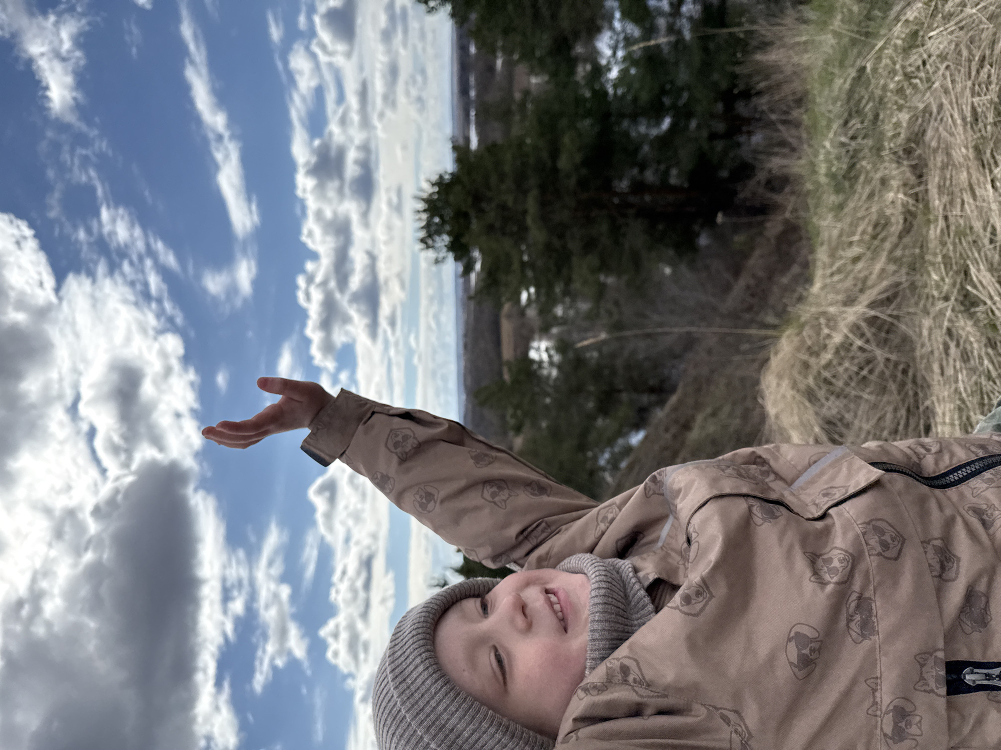 A young child is lying on their side outdoors, wearing a gray knit hat and a light brown jacket patterned with animal faces. The child is smiling and reaching an arm towards the sky. The background includes a blue sky with scattered white clouds, evergreen trees, and dry grass. The location is possibly in a natural area in or near the city of Glazov, Russia, based on image data, although specific landmarks are absent.