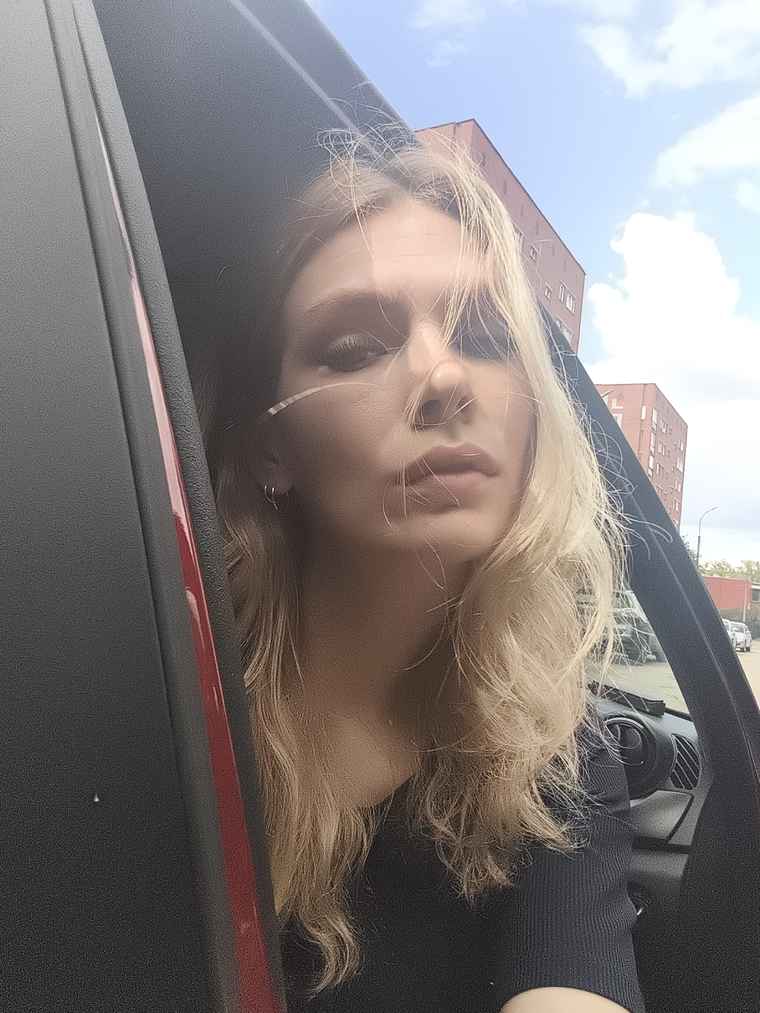 A young woman with blonde hair is captured in a selfie taken from inside a vehicle. She is looking directly at the camera, with her face angled slightly away. Her hair is styled loosely, with some strands falling across her face. She is wearing a dark, ribbed top. The interior of the car is visible, including part of the dashboard and steering wheel. The car door is open, framing the woman's face. Outside the car, there are brick buildings and a blue sky with clouds, suggesting it is daytime and fair weather. The scene appears to be in an urban or suburban environment. The overall mood is casual and candid. There is no visible text in the image.