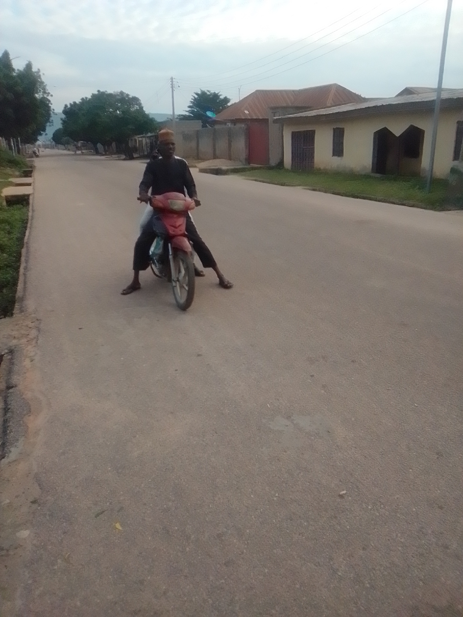 A person wearing a dark top and dark pants, with a cap on their head, is seated on a red motorcycle on a paved road. The person is positioned centrally in the frame, with their legs straddling the motorcycle. The motorcycle is facing towards the viewer. The road appears to be in a residential area, with buildings visible on the right side of the image. Trees line the left side of the road, and a power line runs overhead. The sky is overcast. The image is taken outdoors in Liji Hill, Nigeria.