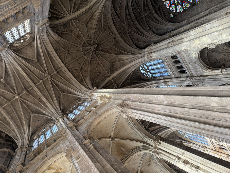 The image displays a high-angle view looking upward within the interior of the Église Saint-Eustache, a Gothic church located in the 1st arrondissement of Paris, France. The scene is dominated by monumental stone architecture, characterized by towering fluted columns, intricate ribbed vaulting, and large windows.
Multiple massive, light-colored stone columns extend vertically into the upper frame, supporting a complex network of pointed arches and elaborate ribbed vaults forming the ceiling. The vaulting patterns vary, featuring both multi-ribbed fan vaults and star-shaped rib designs, culminating in decorative bosses at intersections. Natural light illuminates the interior through numerous tall, arched, multi-pane windows positioned along the upper walls, with the glass appearing clear or faintly tinted blue. In the lower right quadrant, a section of a large circular stained-glass window, possibly a rose window, is visible, featuring various colored glass panes including red, blue, green, and yellow. The stone surfaces exhibit a uniform light brown to grayish hue, consistent with aged masonry. No individuals are present, and no specific actions or interactions are depicted.