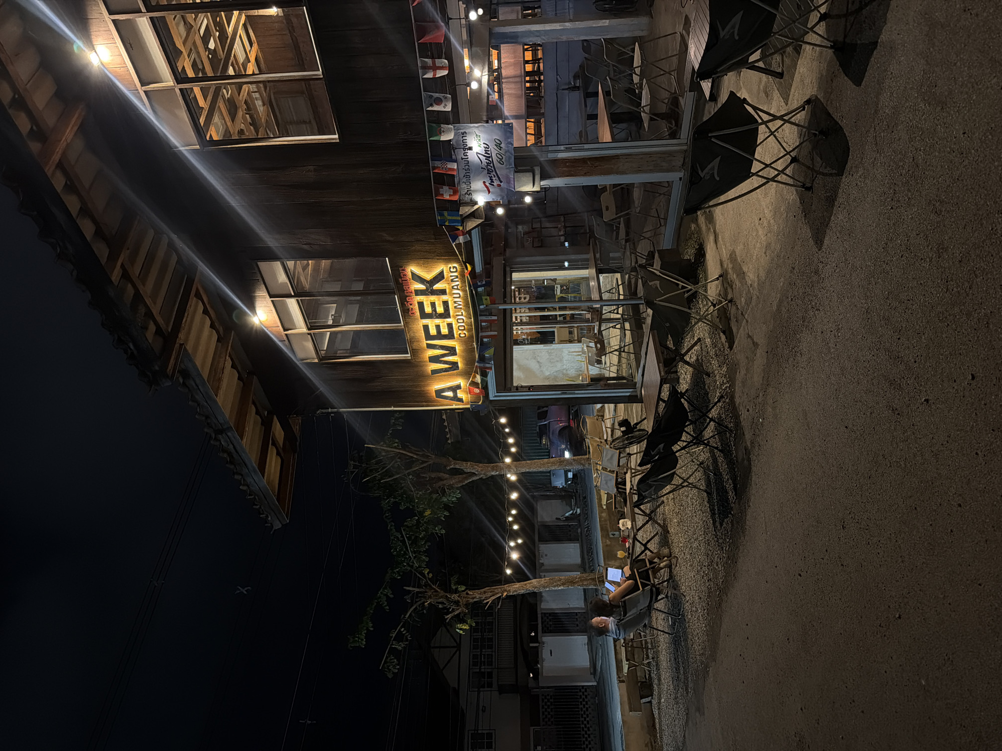 A commercial establishment, identified as "A WEEK COOLMUANG," is depicted at night. The building features a wooden exterior and is illuminated by warm, exterior lights and a prominent, lit sign. String lights are visible, extending across the outdoor seating area. Several tables and chairs are arranged on a gravel and asphalt surface, indicating an outdoor dining or seating space. Two individuals are seated at a table, with one appearing to be using a laptop. The surrounding environment includes trees, streetlights, and neighboring buildings, suggesting an urban or suburban setting. The scene captures a casual evening atmosphere at a commercial establishment.