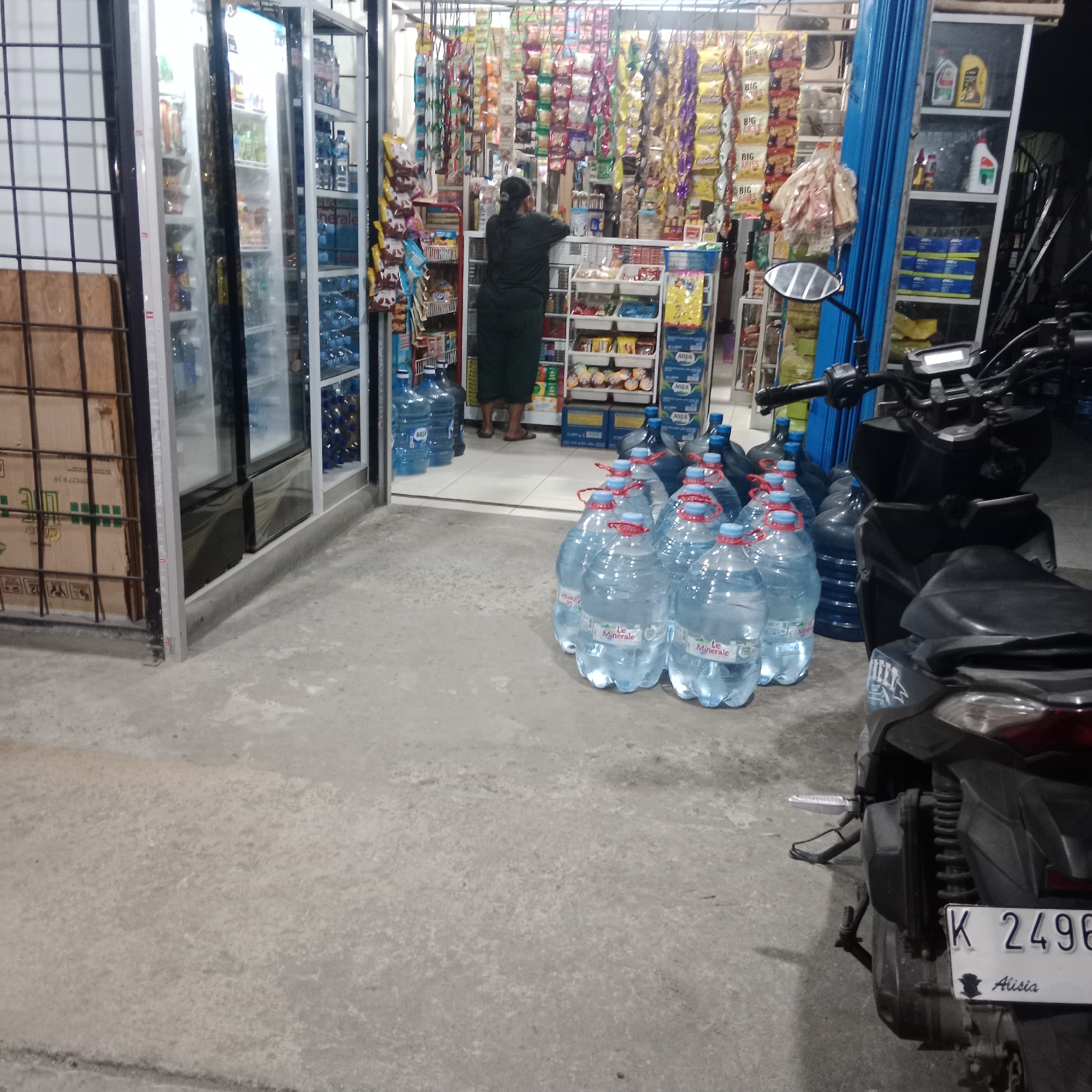 In Gubug, Indonesia, a dimly lit street-side convenience store is visible from the exterior. The store features a glass-fronted refrigerator stocked with beverages and numerous shelves filled with packaged goods and snacks. Outside the store, several large plastic bottles of drinking water are stacked on the concrete ground. A person, dressed in dark clothing, is seen standing inside the store, appearing to be engaged with the merchandise. A black motorcycle is parked in front of the store, with its license plate clearly visible, displaying "K 2496 Alisia." The overall scene suggests a typical small business operation in a semi-urban or rural Indonesian setting.