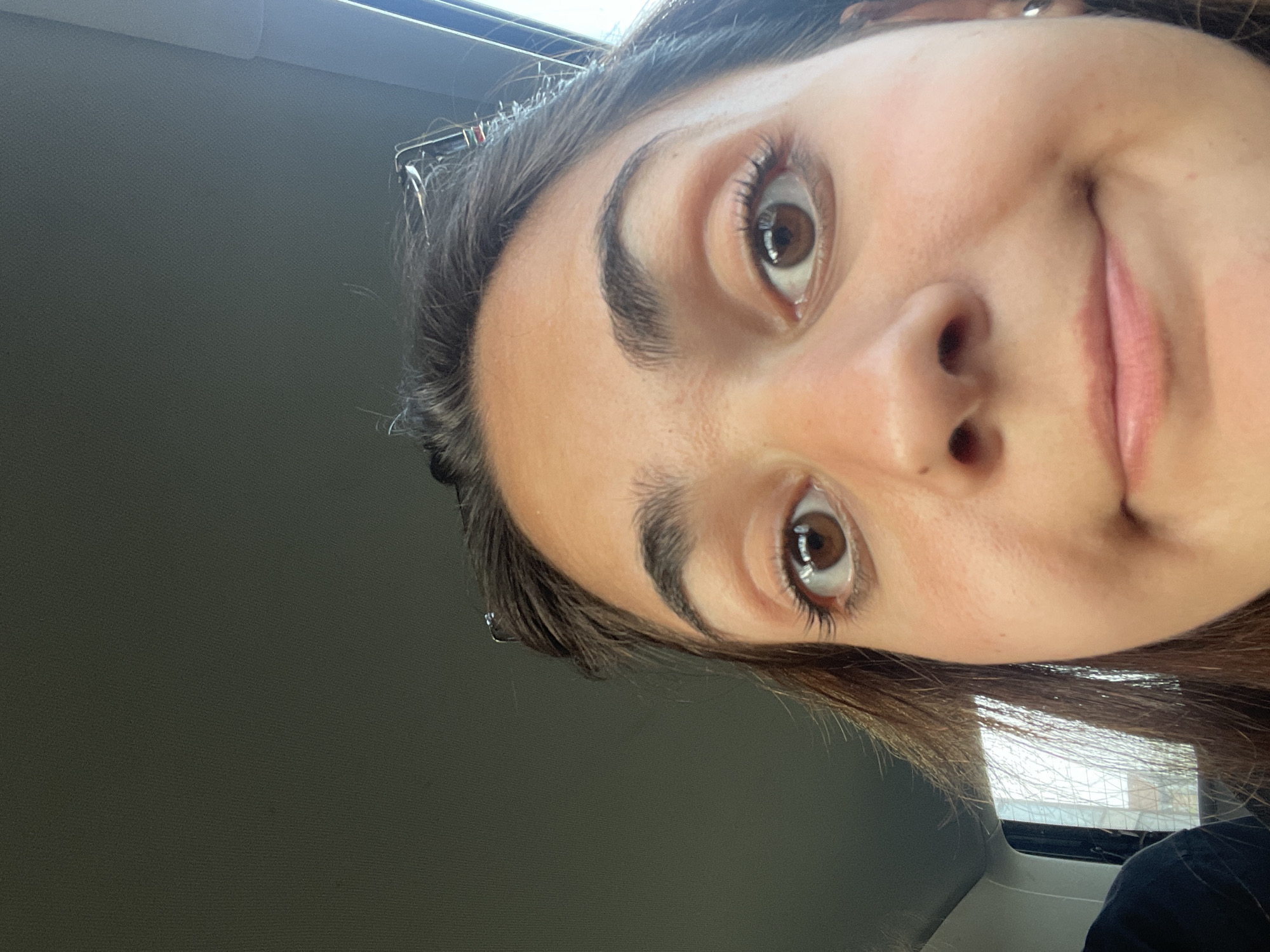 A close-up, downward-angled selfie shows a woman with brown eyes, brown hair, and light skin. Her face fills much of the frame. Part of a gray wall or panel is visible to the left, and a window is partially seen at the bottom right. The location is not discernible from the image.