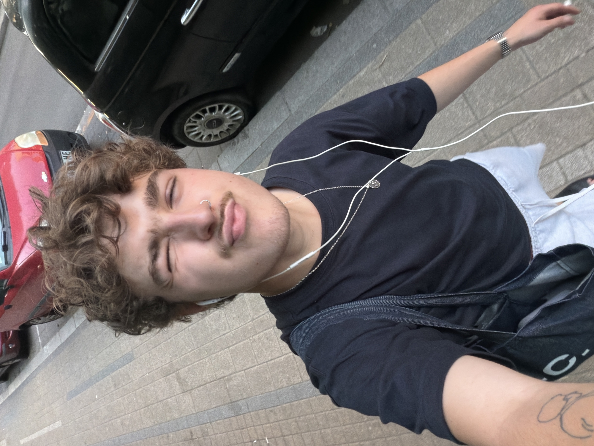 A tilted eye-level selfie depicts a young person on a sidewalk in Vracar Urban Municipality, Serbia. The person has curly hair, closed eyes, a nose ring, and is wearing a black shirt with a silver necklace and headphones. They have a dark denim bag slung across their body and a silver bracelet on their outstretched arm. A red car and a black car are partially visible parked at the curb. The sidewalk is paved with rectangular stone tiles.