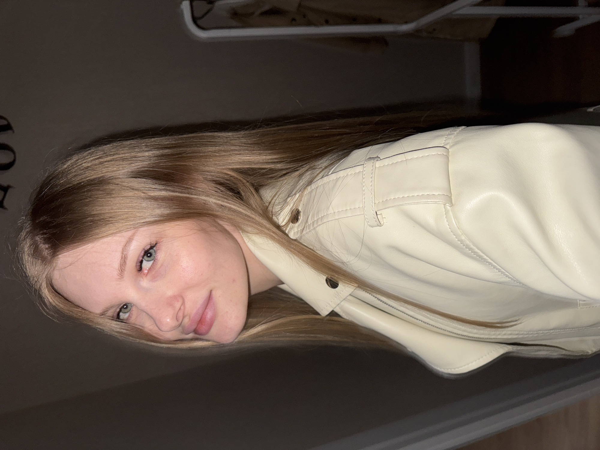 The image displays a head-and-shoulders portrait of a young woman, originally rotated 90 degrees. She has long, straight, light brown hair and fair skin, with light-colored (likely blue or green) eyes. Her expression is calm and friendly, featuring a gentle, slight smile.
The woman is wearing a cream or off-white faux leather jacket, which is a prominent feature of her attire. Notable details on the jacket include visible stitching, a zipper, snap buttons, and a structured design with a subtle quilted-like pattern on the fabric. Her hair falls around her face and over her shoulders, covering parts of the jacket.
The setting is indoors, with a dark and indistinct background, suggesting low lighting conditions. Visible in the background are parts of a white, possibly metal, clothes hanger rack. On a dark wall, there is faint and largely illegible dark lettering or a pattern, with what appears to be part of an "OD" or similar characters visible. The woman is looking directly at the camera, posing for the photograph. The location context is Perm, Russia.