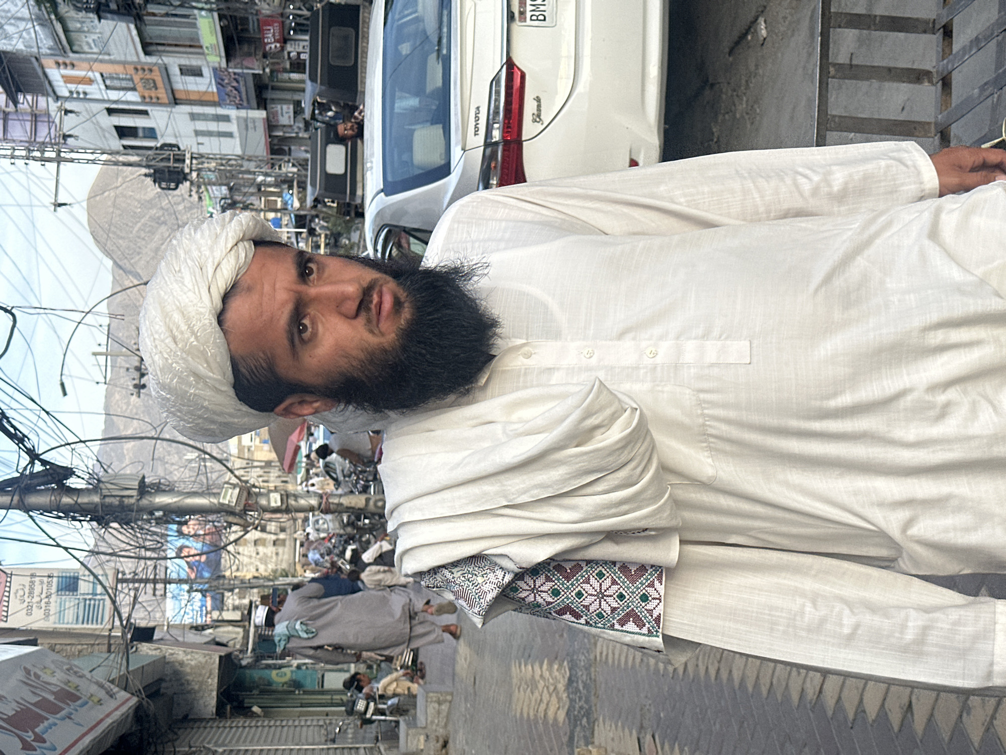 A person in white clothing and turban stands on a sidewalk in Quetta City Tehsil, Pakistan. The person has a dark beard. Behind them is a white car, street wires, and buildings. Other people are walking in the background.