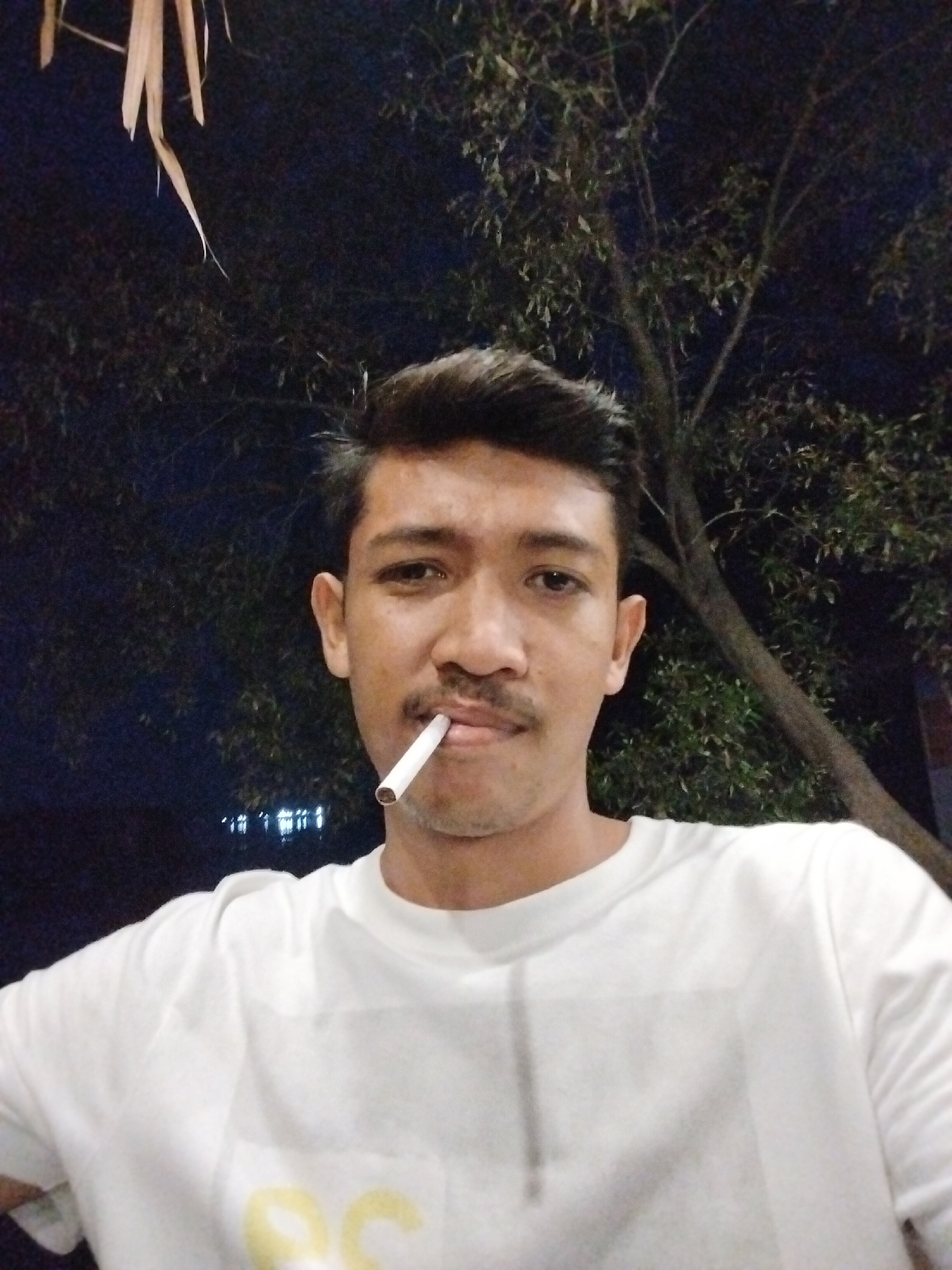 In Lhokseumawe, Indonesia, a young man with short dark hair and a mustache is seen in a close-up, low-angle selfie. He is wearing a white t-shirt. A cigarette is held in his mouth. The background consists of dark foliage and branches of a tree, with faint lights visible in the distance. The overall lighting suggests it is nighttime.