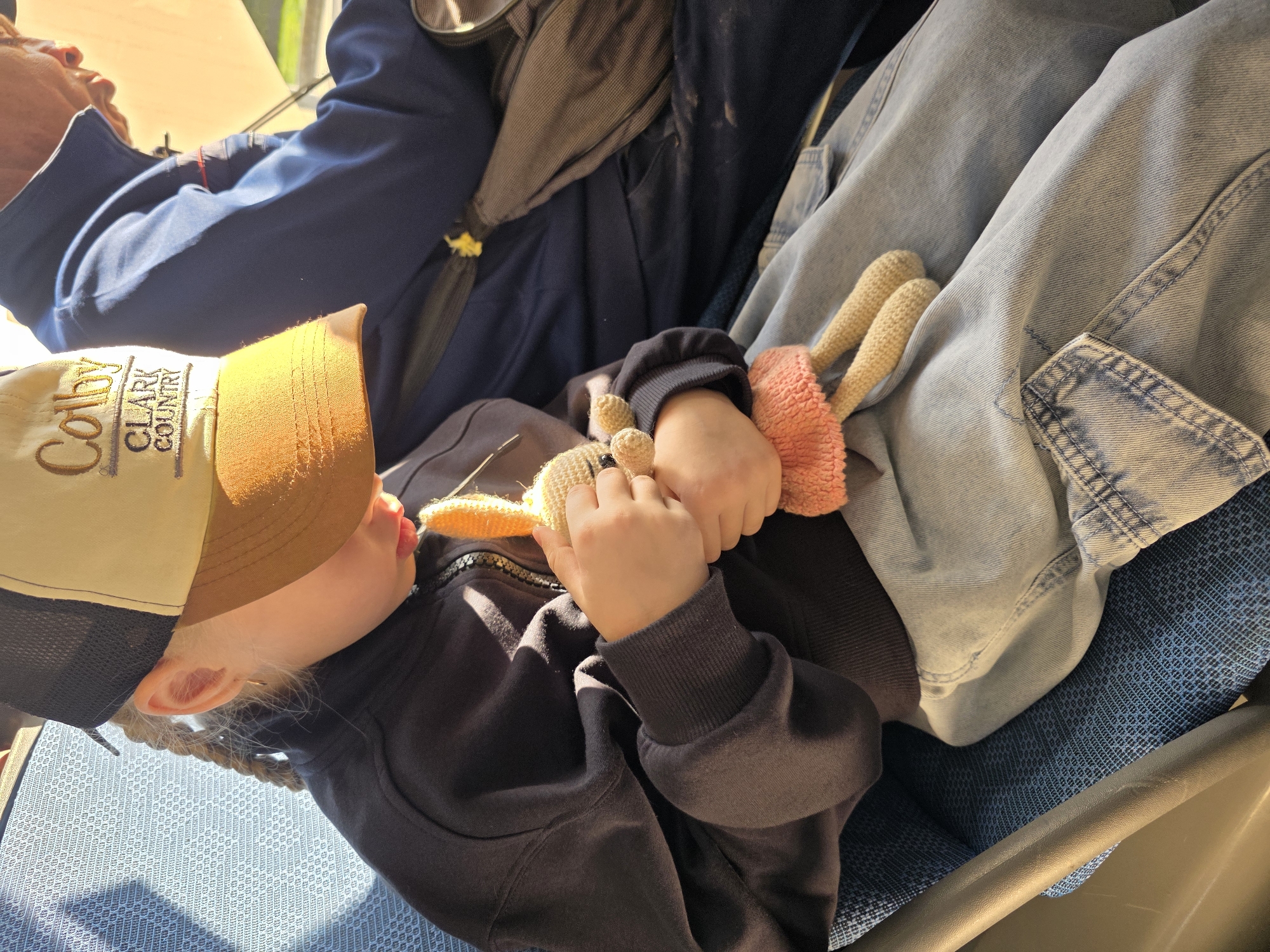 In Kaliningrad, Russia, a young person wearing a dark hooded jacket and a baseball cap with "Colby Clark Country" embroidered on it sits on a blue patterned seat, holding a crocheted doll. The doll has light brown yarn legs and a small pink skirt. The person's light-colored jeans are visible, as are the legs of an adult in a dark blue jacket sitting next to them. The scene appears to be indoors, possibly in a vehicle, with sunlight casting shadows and highlighting the subjects.