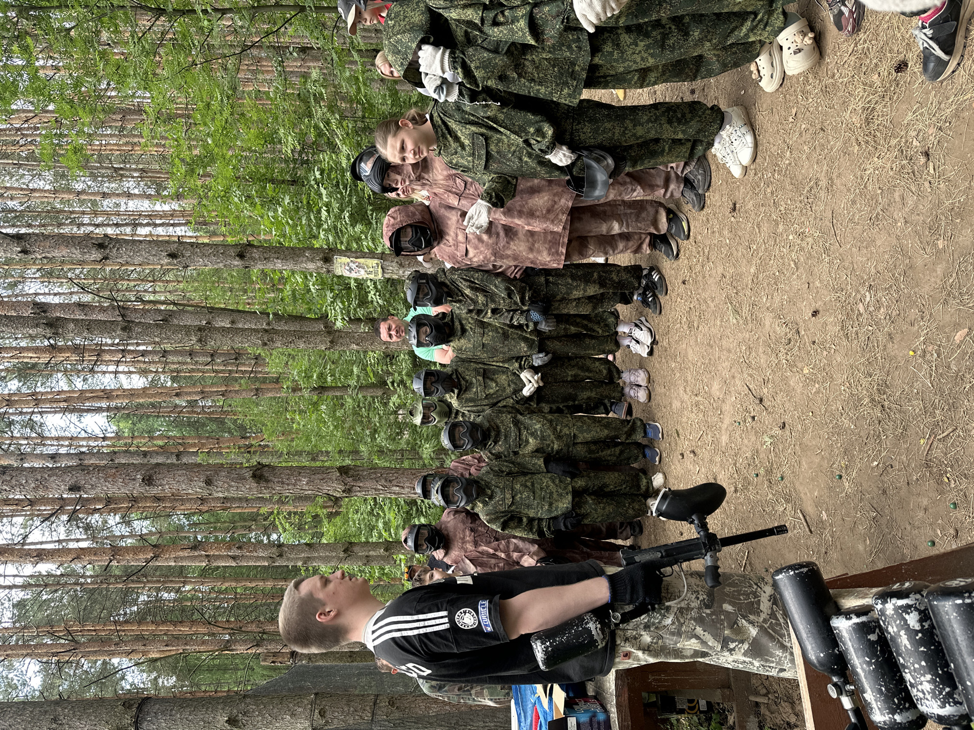 A group of approximately ten people in camouflage clothing and paintball masks stand in a line in a wooded area. One person stands in front of the line holding a paintball gun. Extra canisters of paintballs rest on a wooden bench. The setting appears to be outdoors, possibly a paintball park, with tall pine trees in the background. The location is likely near Votkinsk, Russia.