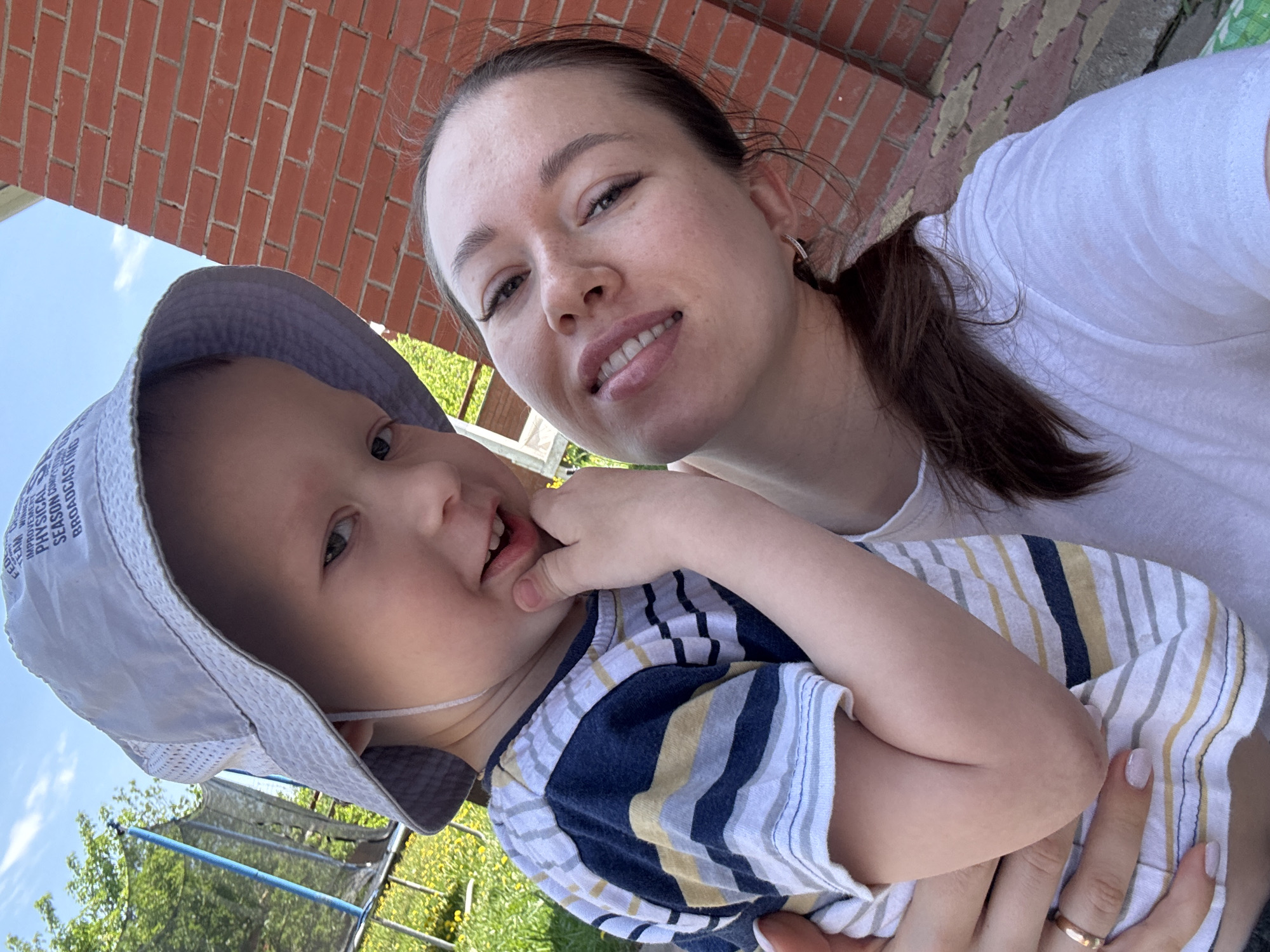 A woman is holding a young child in her arms, likely in an outdoor setting near Ферма, Russia. The woman has brown hair pulled back in a ponytail, fair skin, and is wearing a white t-shirt. She is smiling at the camera. The child is wearing a blue and white striped shirt and a light-colored sun hat. The child has their hand near their mouth. In the background, a red brick wall can be seen. The lower background shows grass, yellow flowers, a green tree and a trampoline. It appears to be a sunny day.