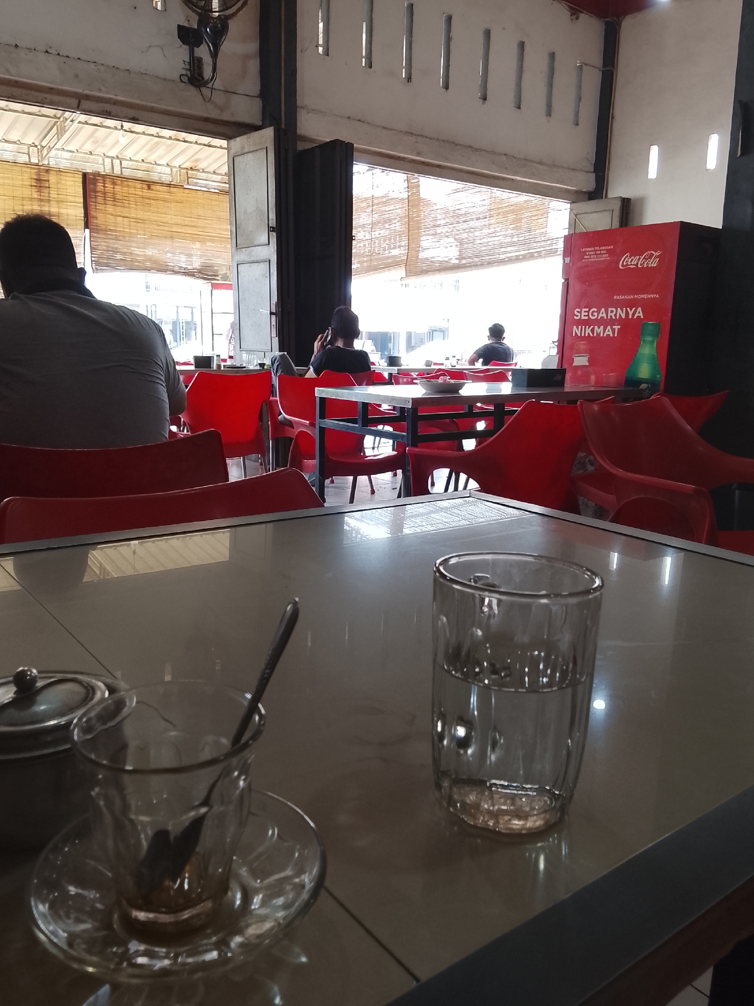 The image shows the interior of a cafe or restaurant, likely in Lhokseumawe, Indonesia.
**Setting:**
The cafe appears to be an open-air establishment with blinds partially covering the windows. The walls are painted white with vertical slots for ventilation. There's a Coca-Cola advertising display in the background.
**People:**
Several people are seated at red tables. One person is facing away from the camera on the left side. In the background, two people are seated at a table, one of whom appears to be looking at a phone.
**Objects:**
In the foreground, a table with a reflective surface holds a glass of water, a small covered container (possibly a creamer), and a glass cup on a saucer with a spoon. The furniture includes red plastic chairs.
**Environment:**
The lighting suggests it's daytime. The cafe setting indicates a place for social gatherings or meals. The general atmosphere appears relaxed and casual.