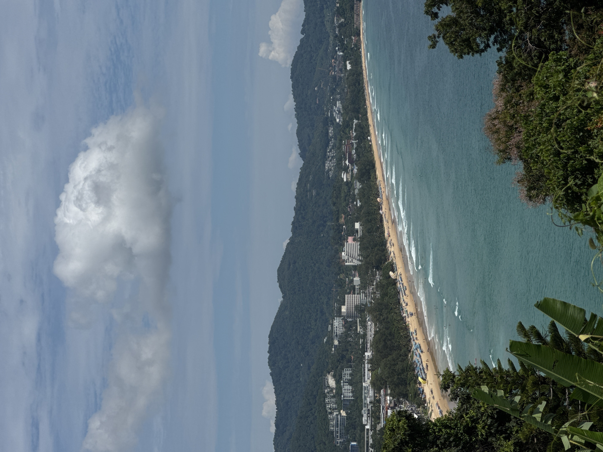 This is a wide shot of Karon Beach in Karon, Thailand. The beach is a long stretch of sand lined with trees and hotels, facing a turquoise sea with moderate waves. A mountain covered in lush green vegetation forms the backdrop to the town. The sky is a bright blue with scattered white clouds. The beach is populated with many people sunbathing, swimming, and walking. Umbrellas and lounge chairs are set up along the sand, and there are some boats in the water.