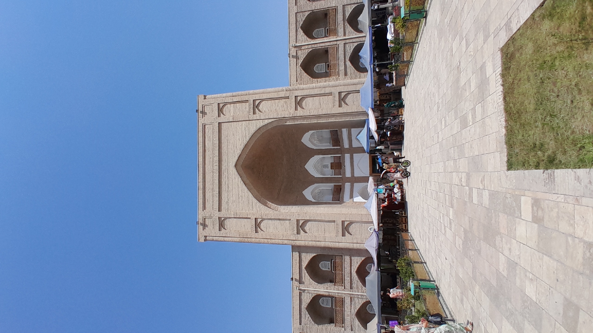 The image depicts the exterior of a large, ancient brick building with an arched entryway, likely a historical site or marketplace in Denov, Uzbekistan. The architecture features several smaller arched windows and decorative brickwork. In front of the building, a paved plaza is set up with numerous market stalls covered by various colored awnings, where vendors appear to be selling goods. Several people are visible browsing the stalls and walking in the plaza. A person is riding a small, motorized scooter-like device through the market. The sky is a clear, bright blue, indicating a sunny day. The ground is paved with large, light-colored rectangular stones, and there is a patch of green grass to the right of the plaza.