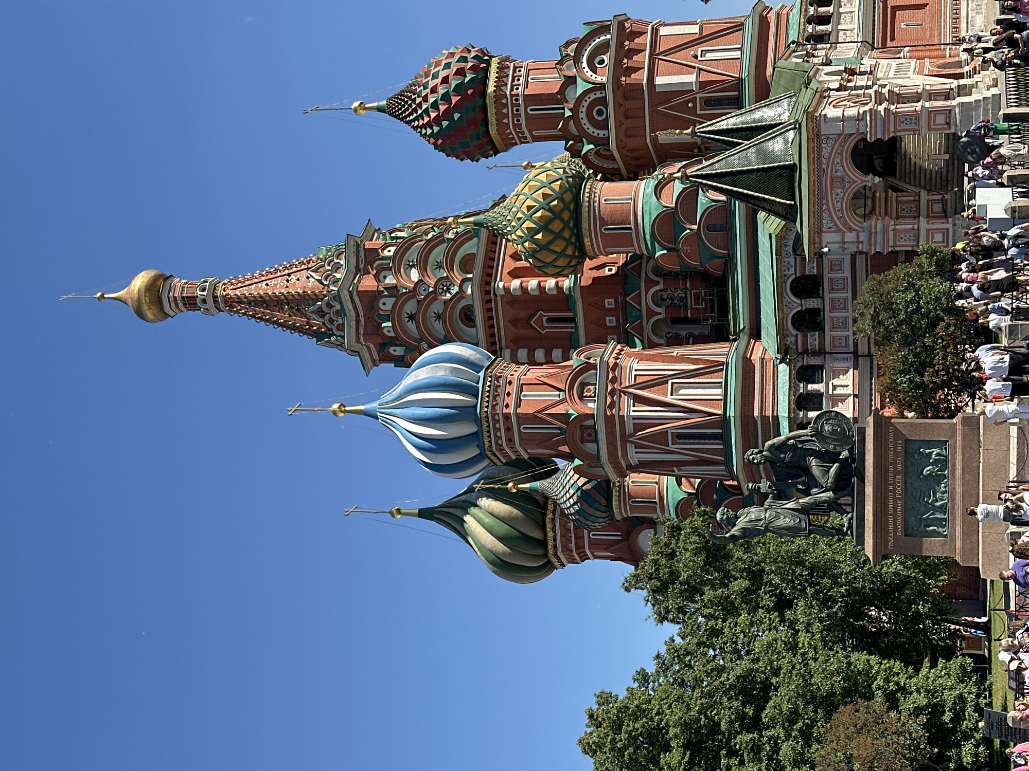 The image depicts St. Basil's Cathedral, located on Red Square in Moscow, Russia, under a clear blue sky. The cathedral features prominent red brick construction with intricate white and green architectural detailing. Numerous onion-shaped domes rise from its structure; visible examples include a central tall golden dome with a cross finial, a large blue and white striped dome, a large green and white striped dome, a smaller green and yellow patterned dome, and a smaller red and green pyramidal patterned dome, each topped with a cross. Green roofs and accents are visible below the domes and along the lower sections of the building.
In the lower-left foreground, the Monument to Minin and Pozharsky stands on a multi-tiered stone pedestal. The bronze sculpture portrays two male figures, one standing with an outstretched arm and the other seated, holding a shield. The pedestal bears an inscription in Cyrillic and a bas-relief panel depicting several figures. Lush green trees partially obscure the base of the cathedral and the background behind the monument.
Numerous individuals, appearing to be tourists, populate the lower portion of the scene, gathered around the monument and near a multi-tiered arched entrance to the cathedral on the right. The lighting indicates a bright, sunny day, with the sun casting clear illumination on the subjects.