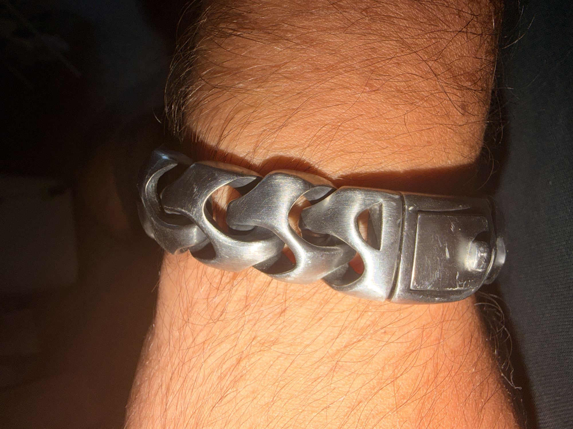 A close-up, eye-level shot shows a thick, silver-colored metal bracelet with large, interlocking links worn on a person's wrist. The bracelet is positioned horizontally across the frame, with the clasp visible on the left side. The wrist is covered in fair skin with visible hair. The background is dark and out of focus, suggesting an indoor setting. There is no indication of the location being Castillo Bajo-conejito, Spain.