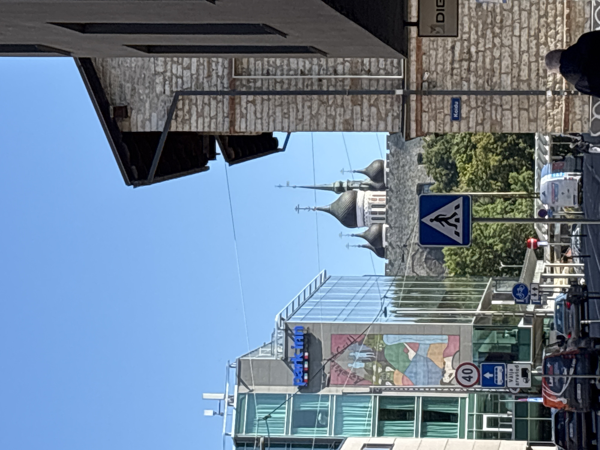 This image captures a street scene in Tallinn, Estonia, on a clear day. In the foreground, a modern building with a glass facade, displaying the "Park Inn" logo, stands to the left. Adjacent to it, a traditional stone building occupies the right side of the frame. Beyond these buildings, the distinctive onion domes of a Russian Orthodox cathedral are visible, suggesting a prominent landmark in the city. Several street signs are present, including a pedestrian crossing sign, a speed limit sign indicating 40, and a directional sign with a bus symbol and text that appears to be in Estonian. Cars and vans are on the street, with a pedestrian walking away from the camera in the lower right corner. The sky is a clear, bright blue, indicating fair weather.