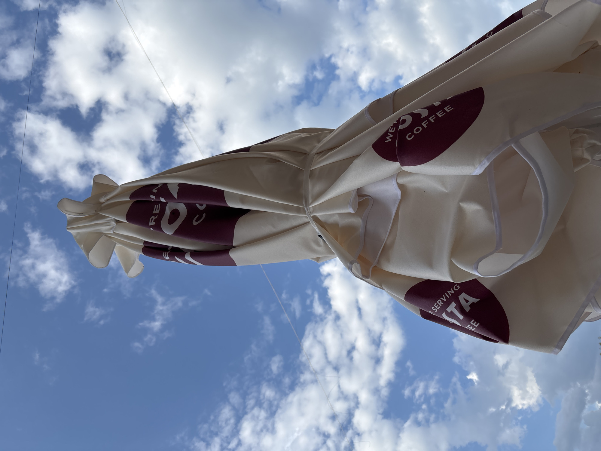 The image, taken from a low angle, showcases a closed Costa Coffee umbrella against a backdrop of a blue sky dotted with fluffy white clouds. The umbrella, predominantly white with burgundy branding ("WE'RE SERVING COSTA COFFEE"), is secured with a white tie. Fine lines, possibly wires, are visible against the sky in the upper portion of the frame. The location is the city of Garmen, Bulgaria.