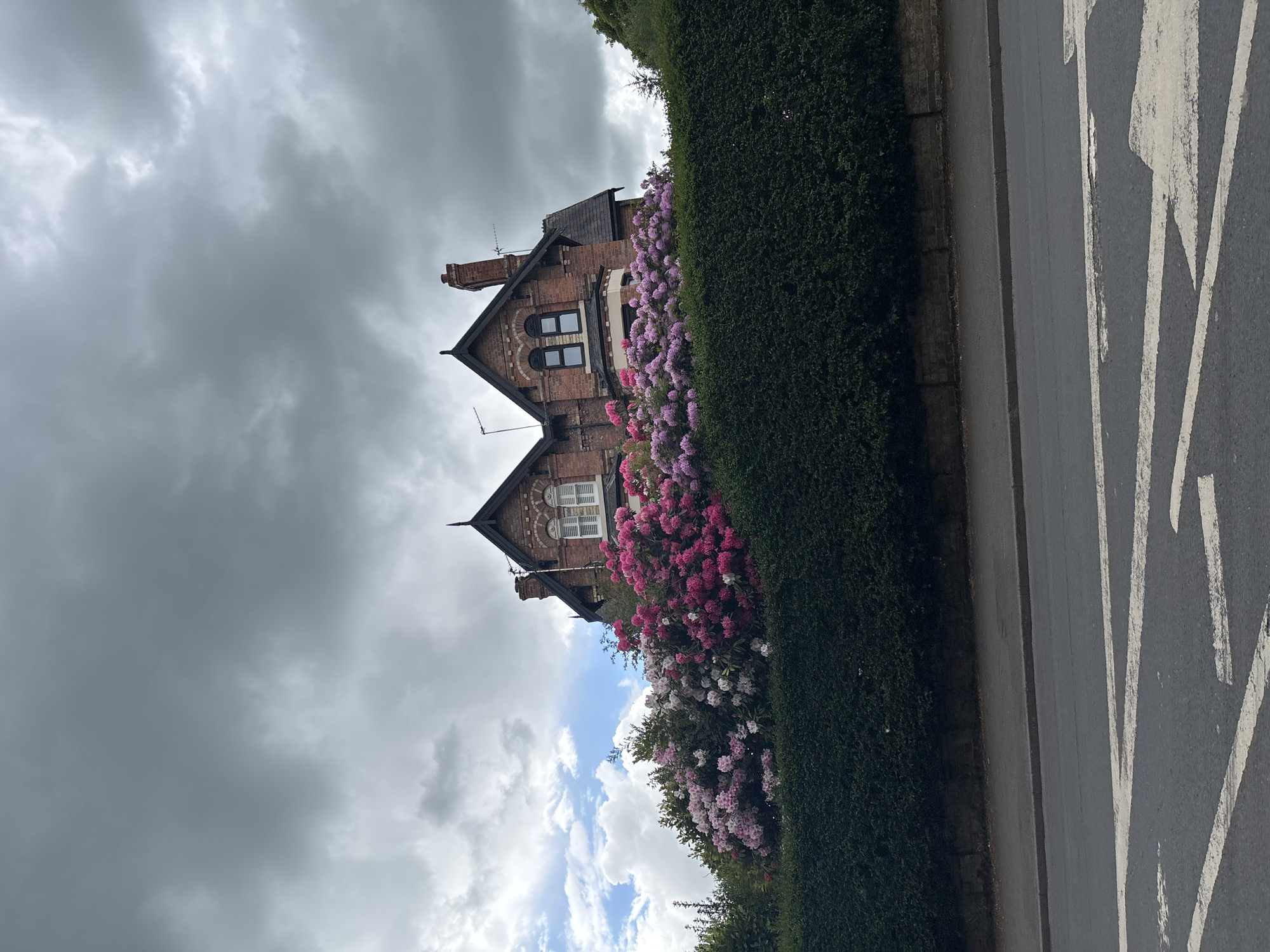 The image shows a residential street in Knutsford, United Kingdom. A two-story brick house with a steeply pitched roof is visible. The house is partially obscured by a tall, manicured hedge adorned with blooming rhododendrons in shades of pink and purple. The sky is overcast with dark, looming clouds, but patches of blue sky are visible behind the rhododendrons. To the right of the hedge is an asphalt road with white painted road markings. There are no people visible in the scene.