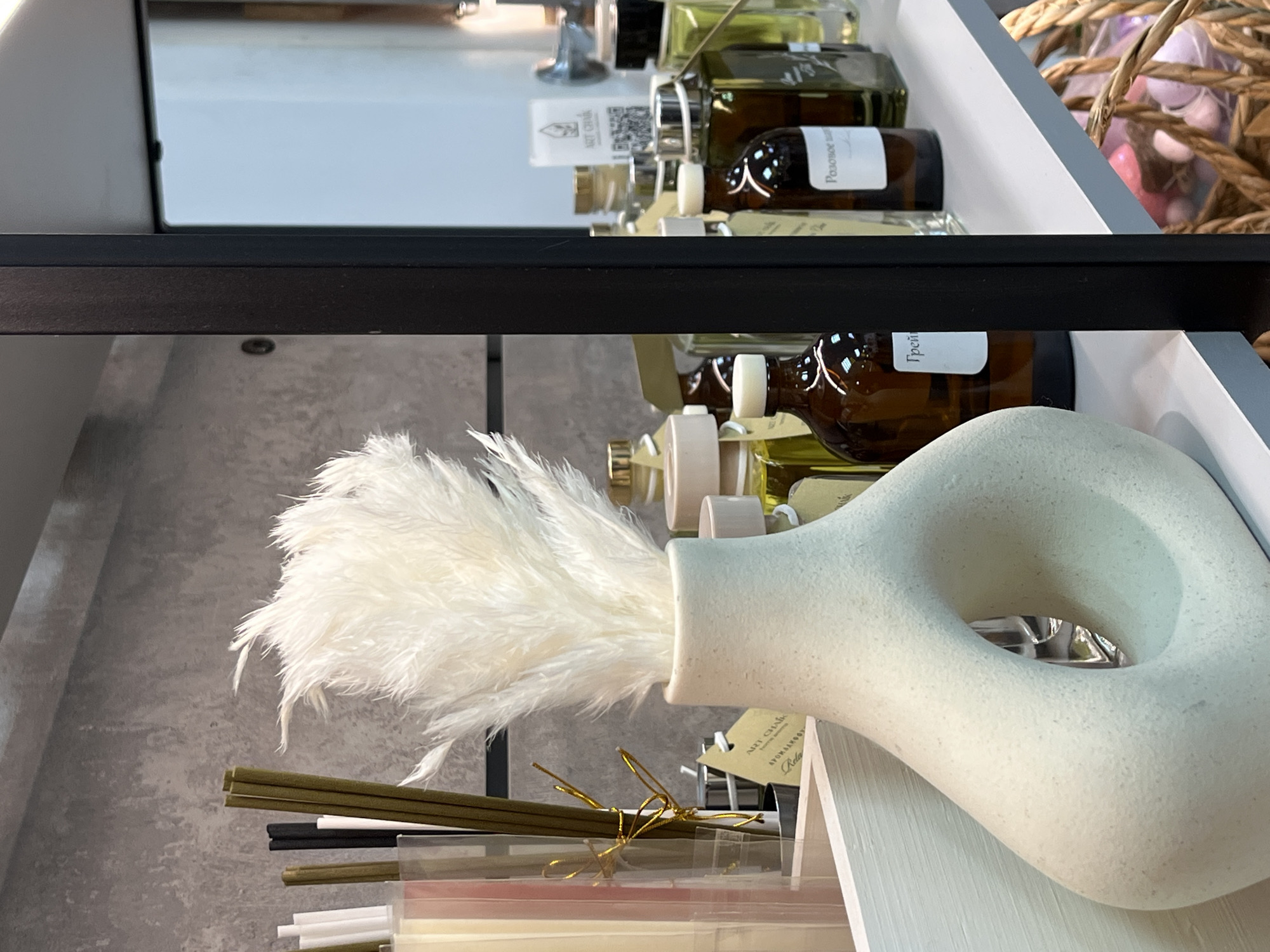 The image depicts an interior setting, showcasing a shelf displaying various items, potentially in a shop or a retail environment. The focal point is a white, donut-shaped vase with feathery plumes arranged in it. Below the vase, there are bundles of sticks or reeds, likely part of a decorative arrangement or product.
Above the vase and reeds, the shelf holds an assortment of glass bottles filled with liquids, suggesting they could be perfumes, essential oils, or similar products. The bottles have labels, some of which are partially visible. One label includes a QR code and the text "ART CHAIK," indicating that the place could be the city of Chaikovsky, Russia.
The shelf is enclosed by a frame, with a black bar running across the image's midsection. The background consists of textured, grey surfaces, which could be walls or additional shelving units. The overall arrangement is neatly organized, suggesting a focus on aesthetic presentation for potential customers.